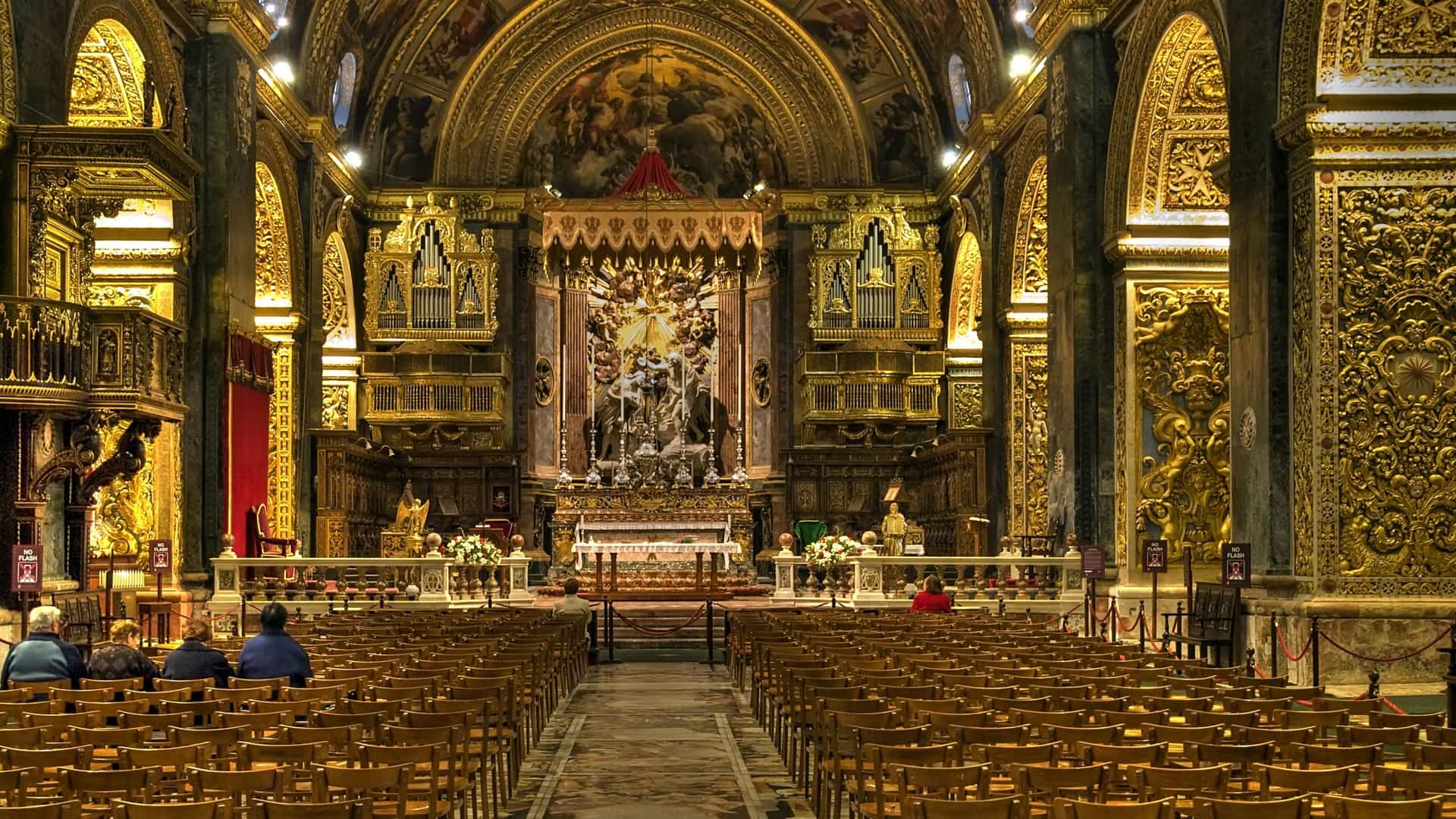 The opulent, Baroque interior of St. John's Co Cathedral in Valletta, Malta. The view focuses on the richly decorated vaulted ceiling and the side aisles. The entire space is lavishly adorned with marble tombstones covering the floor, intricate gold leaf ornamentation, and detailed masonry, all illuminated in a high dynamic range (HDR) photograph.