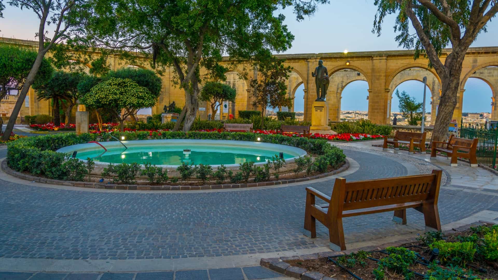 A sunset view of the Upper Barrakka Gardens in Valletta, Malta, highlighting the ornate stone architecture. The image features a prominent arcade of decorative limestone arches and columns, framed by palm trees and lush plants, with the warm light of the setting sun casting long shadows. The scene captures the picturesque viewpoint over the city and waterfront.