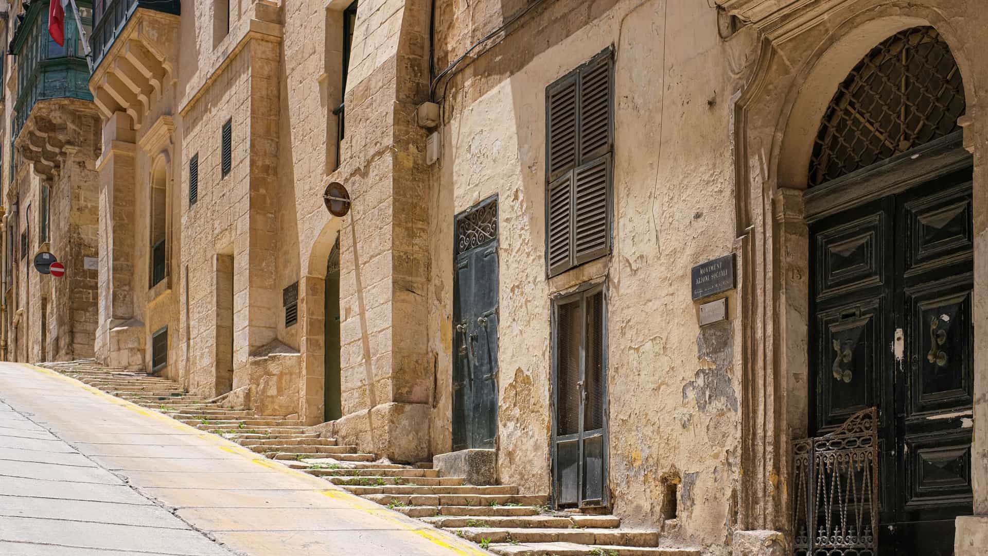 A view looking up a narrow, steep, hilly street in the historic city of Valletta, Malta. The street is lined with traditional limestone buildings that feature colorful, enclosed wooden balconies known as gallarija.