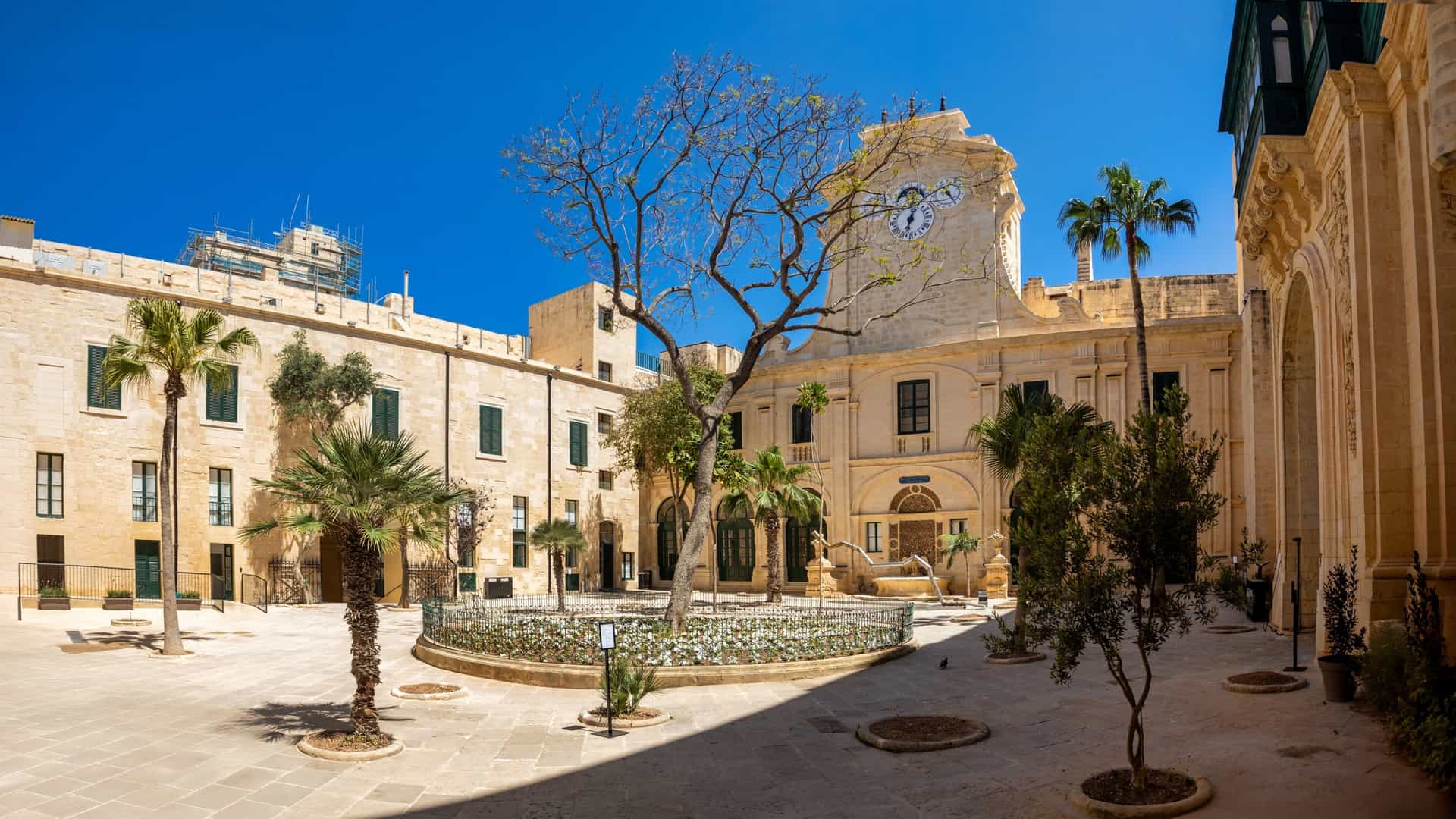 A view of a historic courtyard within the Grandmaster's Palace in Valletta, Malta. The courtyard is surrounded by two story architectural facades of light colored stone, featuring covered wooden balconies (gallarija) and open arched loggias on the second floor. In the center is a statue of Neptune on a pedestal, with surrounding green plants and trees.