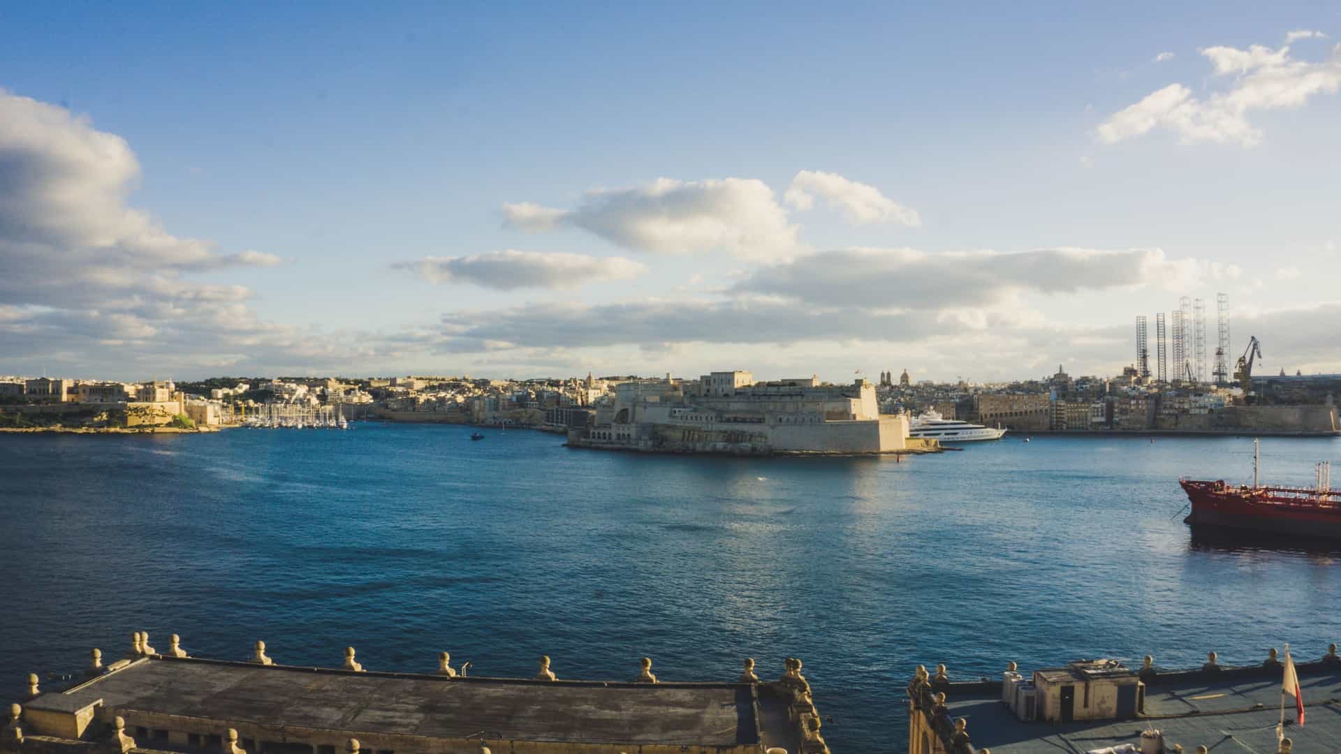 A bright, panoramic view from the Lower Barrakka Gardens in Valletta, Malta, overlooking the Grand Harbour. In the foreground, the neoclassical Monument to Sir Alexander Ball, a circular temple with Doric columns and a domed roof, is visible among lush green trees and manicured gardens. In the background, the Grand Harbour is visible, with the Valletta Breakwater and the Mediterranean Sea extending to the horizon under a clear sky.