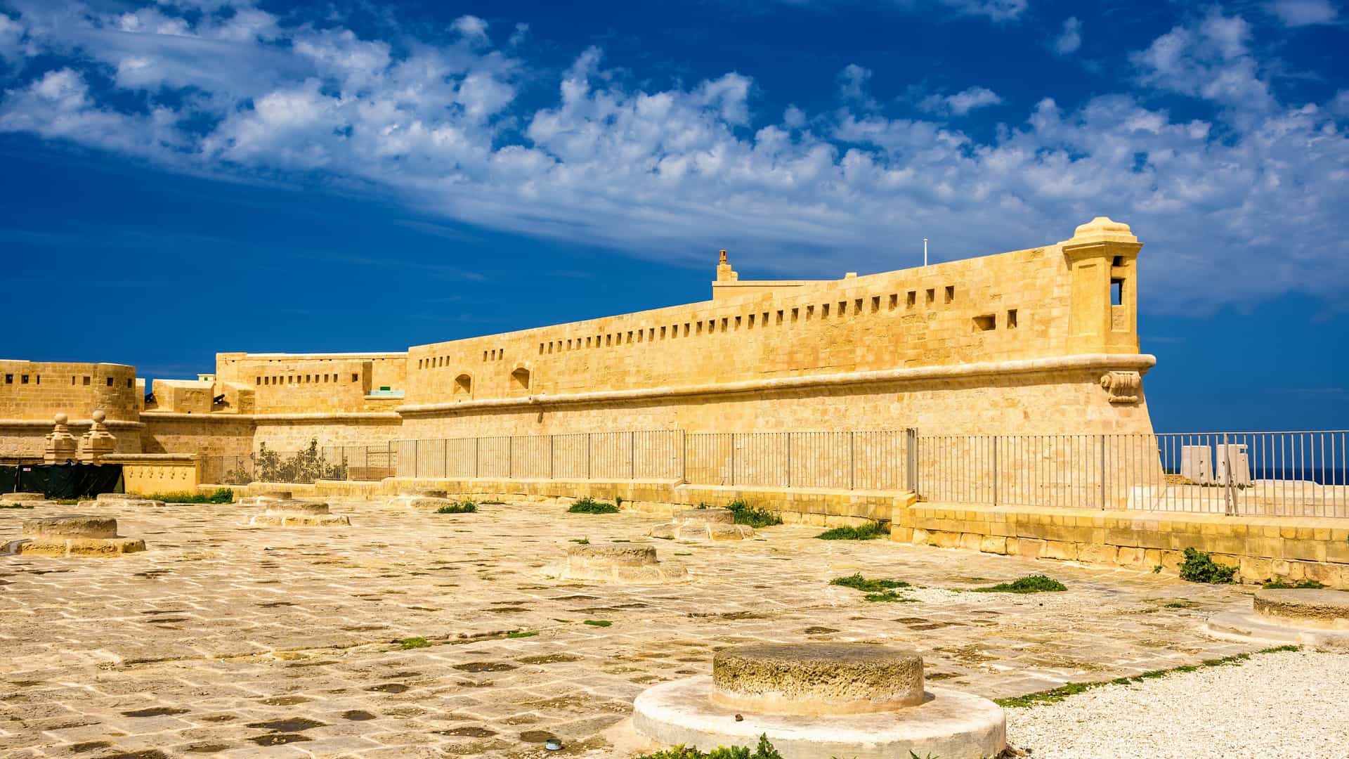 An exterior view of Fort Saint Elmo, a star shaped fortification located at the tip of the Sciberras Peninsula in Valletta, Malta. The massive stone walls, bastions, and ramparts of the historic fort are visible, overlooking the Mediterranean Sea. The fort's construction shows its role as a key defensive structure protecting the entrances to the Grand Harbour and Marsamxett Harbour.