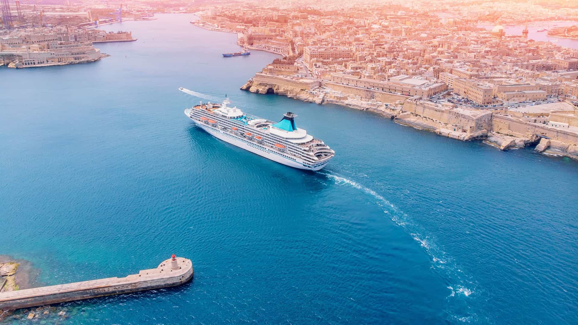 An aerial, high angle view of a large cruise ship docked in the Grand Harbour of Valletta, Malta. The massive white liner is positioned in the port, with the historic, fortified city's skyline of yellow stone Baroque buildings and bastions visible on the surrounding hillsides.