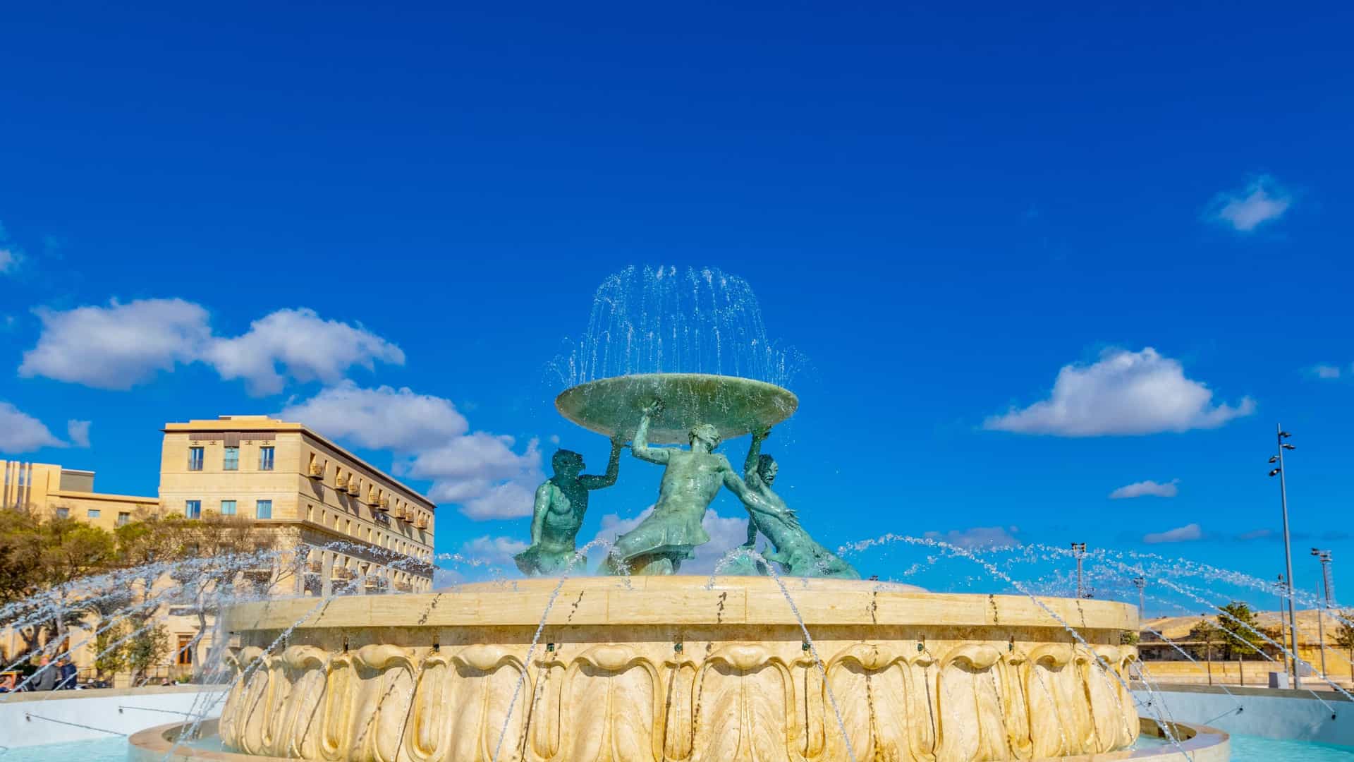 A photo of the famous Triton Fountain located just outside the City Gate of Valletta, Malta. The centerpiece consists of three massive bronze figures of mythological Tritons, balanced on a seaweed like base, holding up a large circular basin. Water jets spray dramatically from the base and the basin, and in the background, the modern Parliament House and the entrance to Valletta are visible.