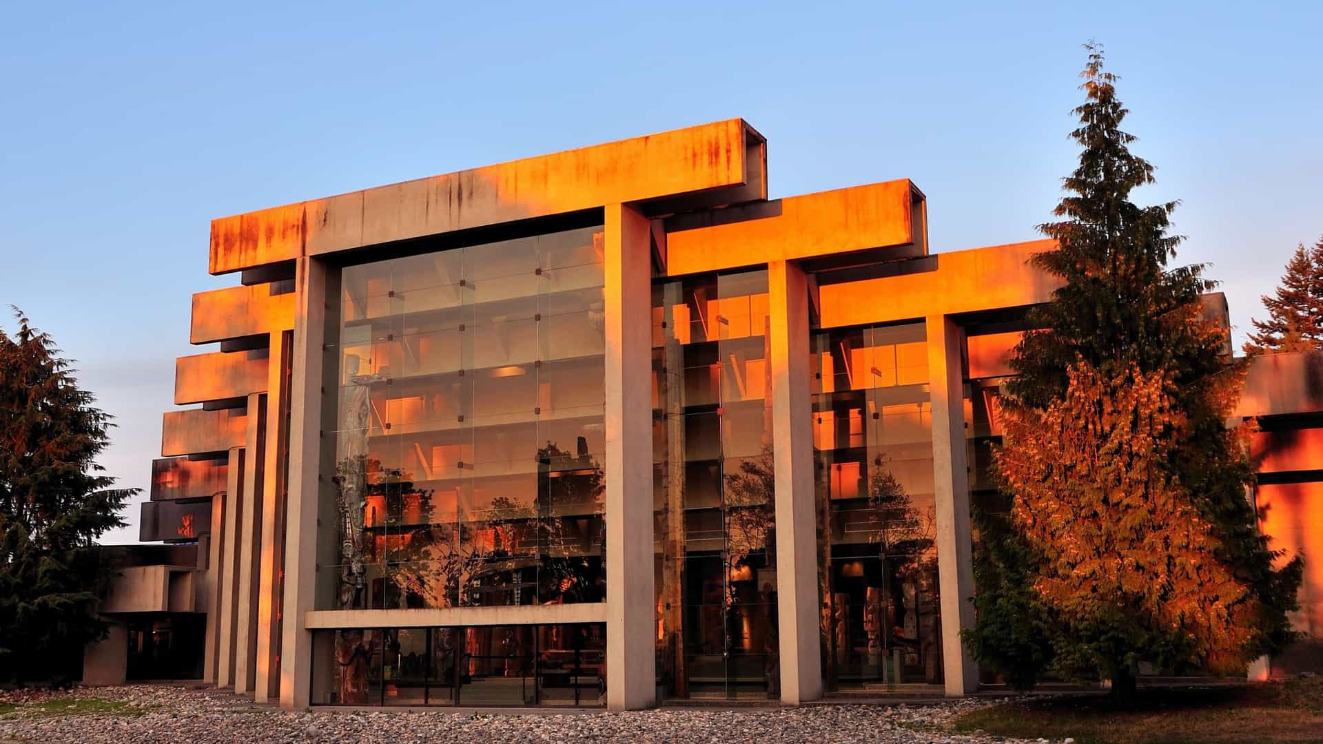 The modern, angular concrete and glass architecture of the Museum of Anthropology at the University of British Columbia (UBC), with several towering First Nations totem poles standing prominently on the museum grounds outside.