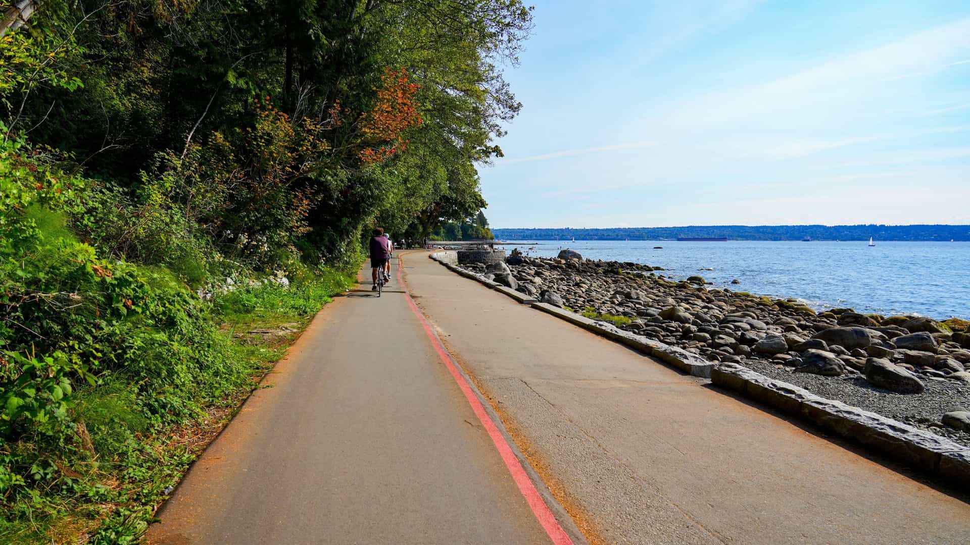 The Vancouver Seawall pedestrian and cycling path along the coast of English Bay, with a low stone barrier separating the paved path from the water. The image captures the urban waterfront with the calm bay on one side and the modern high rise buildings of the city's West End neighborhood on the other, all under a clear sky.
