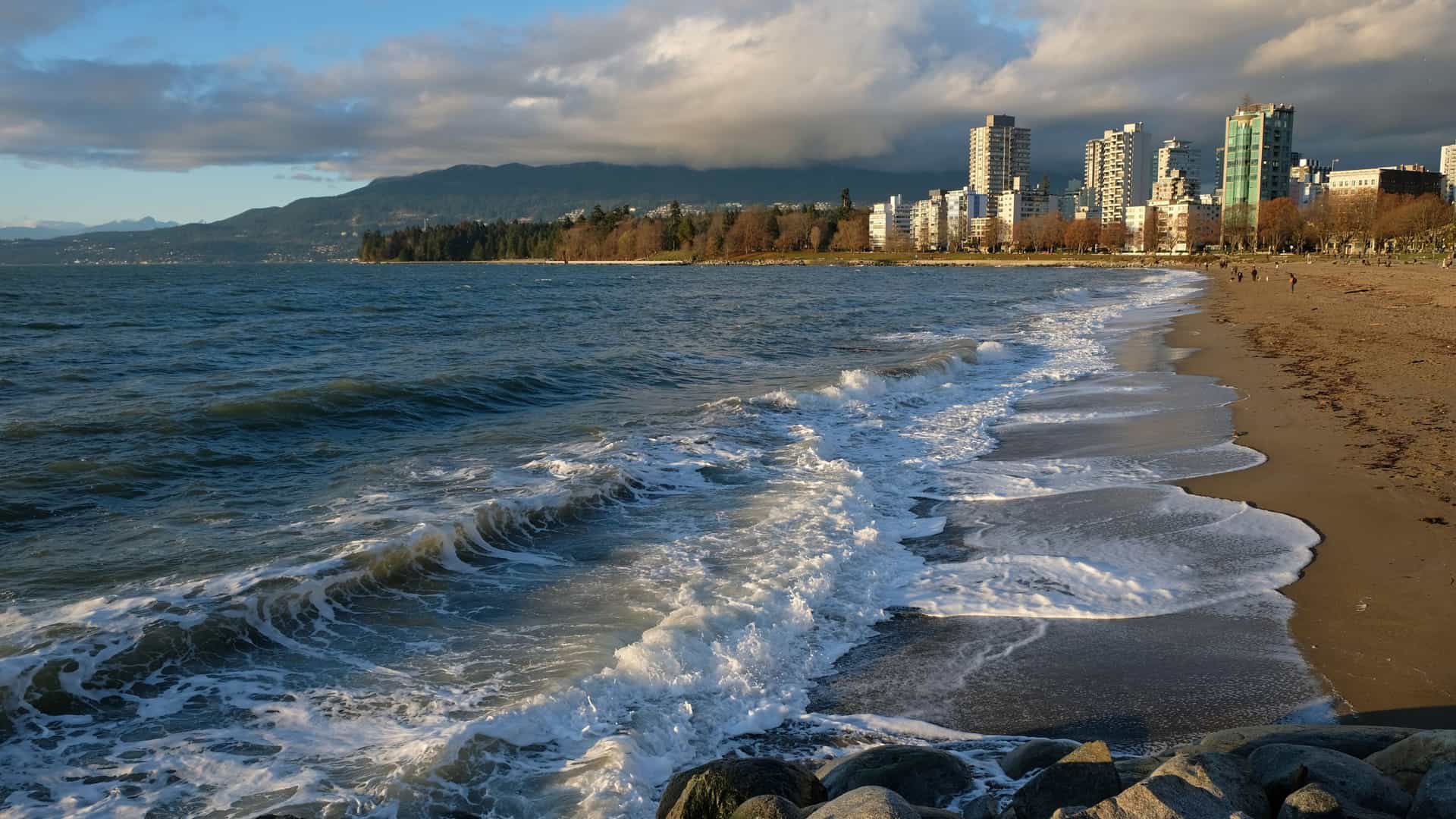 Close up shot of moderate waves washing up onto the sandy shore of English Bay Beach in Vancouver, British Columbia, with the foam from the breaking waves visible on the wet sand. The water's surface shows a mix of blue and white, and the background is slightly blurred but suggests a view of the bay.