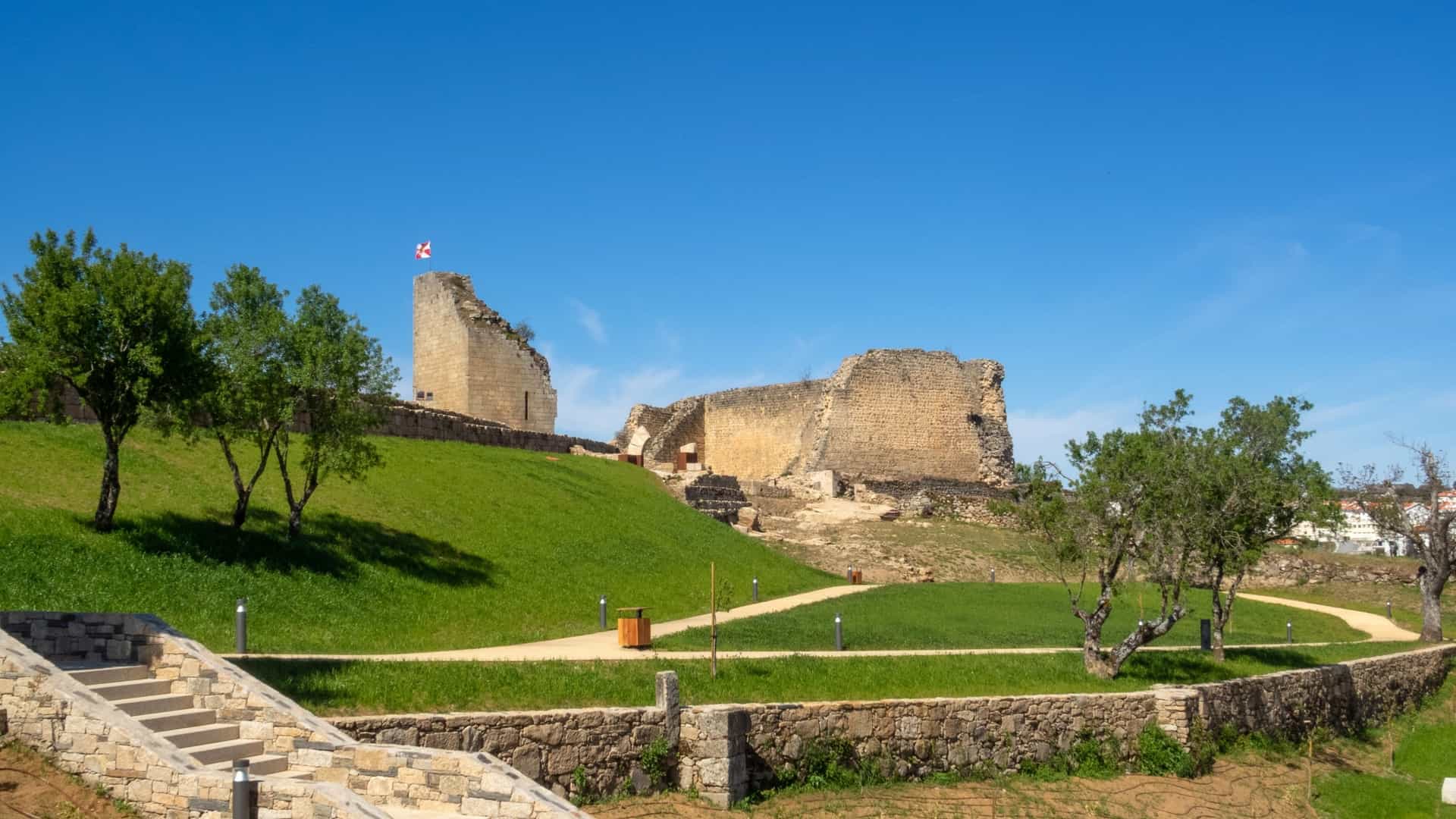A scenic, wide angle photo of the ruins of the Miranda do Douro Castle, located on a rocky cliff overlooking the Douro River on the border of Portugal (Trás os Montes region). The surviving walls and tower stand against a vast, bright blue sky with a few clouds, conveying the site's historic and strategic significance.
