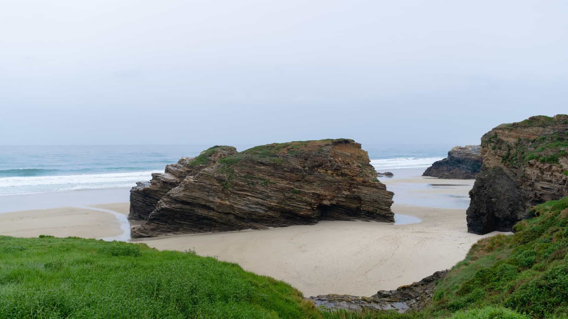 A breathtaking panoramic view of the Playa de las Catedrales (Beach of the Cathedrals) in Ribadeo, Galicia, Spain. The Atlantic Ocean's low tide reveals a vast expanse of sandy beach, intricate sea caves, and towering natural rock arches and cliffs that resemble the flying buttresses of a Gothic cathedral. A person is visible in the distance, providing a sense of scale to the majestic rock formations.