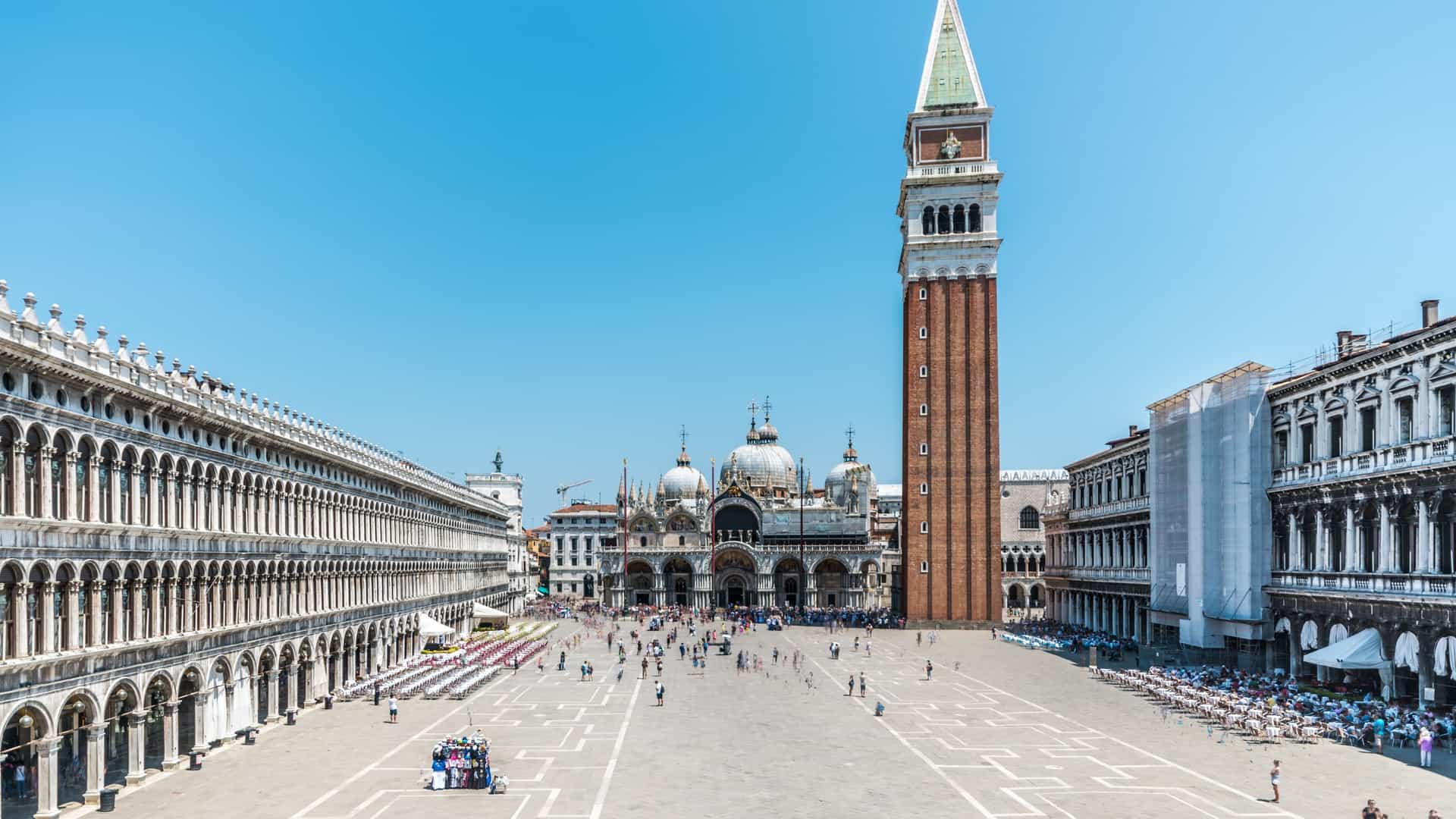 A panoramic view of Piazza San Marco (St. Mark's Square) in Venice, Italy. The iconic square is framed by the arcaded Procuratie buildings, with the ornate, domed facade of St Mark's Basilica and the tall, separate Campanile (bell tower) standing prominently in the background. The square is filled with people and the famous geometric paving.
