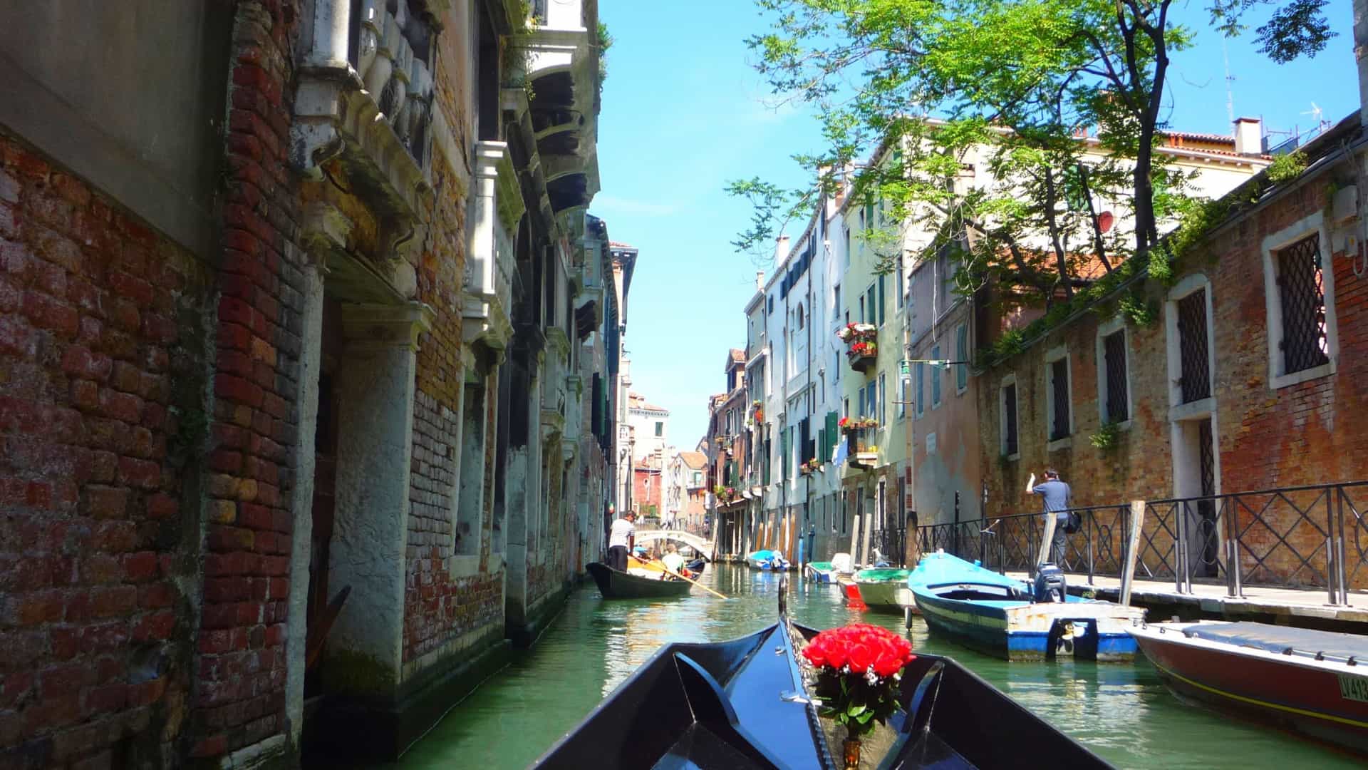 A traditional black gondola carrying tourists is being expertly steered by a gondolier through a narrow, sunlit canal in Venice, Italy. The gondola is flanked by the towering, colorful historic buildings with aged plaster and shuttered windows. The water's surface reflects the architecture and sky.