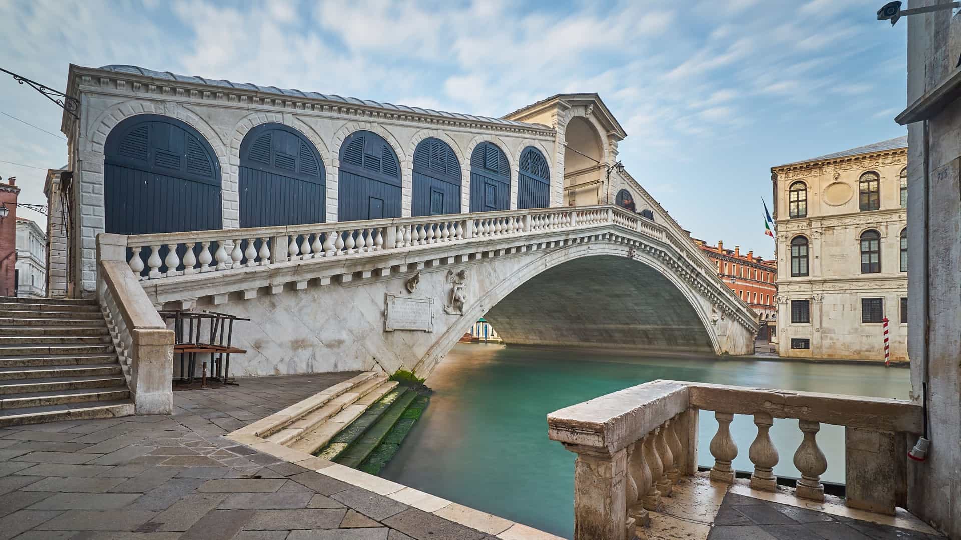 A scenic view of the Rialto Bridge (Ponte di Rialto) spanning the Grand Canal in Venice, Italy, on a clear morning. The white stone arch of the bridge is centered, with historic buildings lining the canal on both sides. A few colorful boats and black gondolas are visible on the calm, reflective water.
