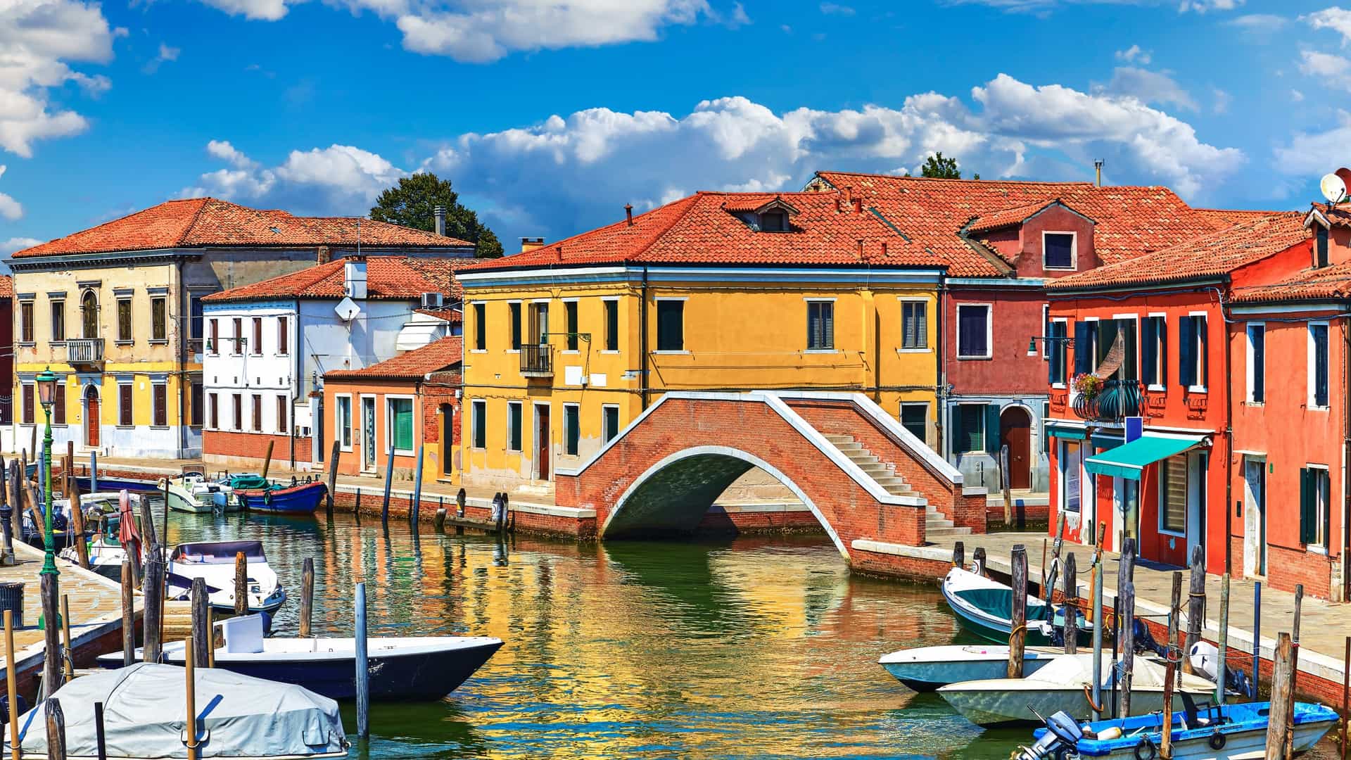 A scenic view of a canal on the island of Murano, near Venice, Italy, on a sunny day. The canal is lined on both sides by traditional buildings with colorful facades in shades of pink, orange, yellow, and red. Several boats and small motorboats are moored along the waterfront. In the distance, the prominent bell tower of a church, likely the Basilica dei Santi Maria e Donato, rises above the rooftops. Murano is famous for its historic glass making industry.