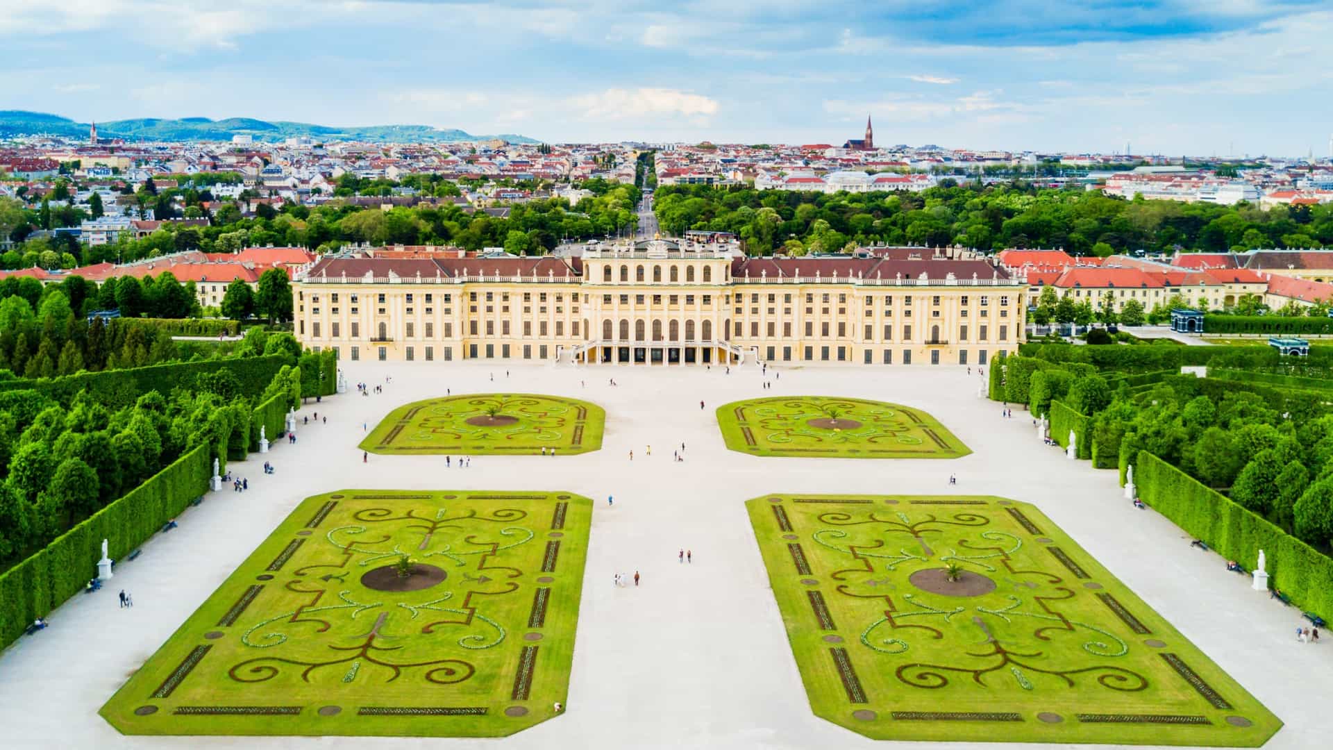 A high angle panoramic aerial view of Schönbrunn Palace (Schloss Schönbrunn) in Vienna, Austria. The yellow Baroque palace is prominently featured, surrounded by its expansive, meticulously manicured formal gardens and fountains, with the cityscape of Vienna visible in the distance.