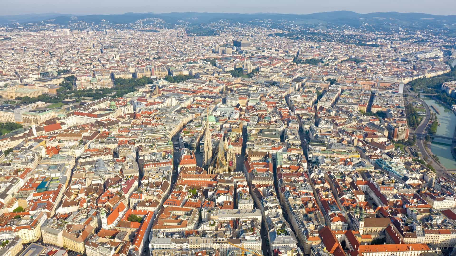 An aerial panoramic view of the historic city center of Vienna, Austria. The cityscape is dense with classical European buildings featuring terracotta and tiled rooftops, with the prominent spire and ornate, colorful patterned roof of St. Stephen's Cathedral (Stephansdom) rising above the urban fabric.