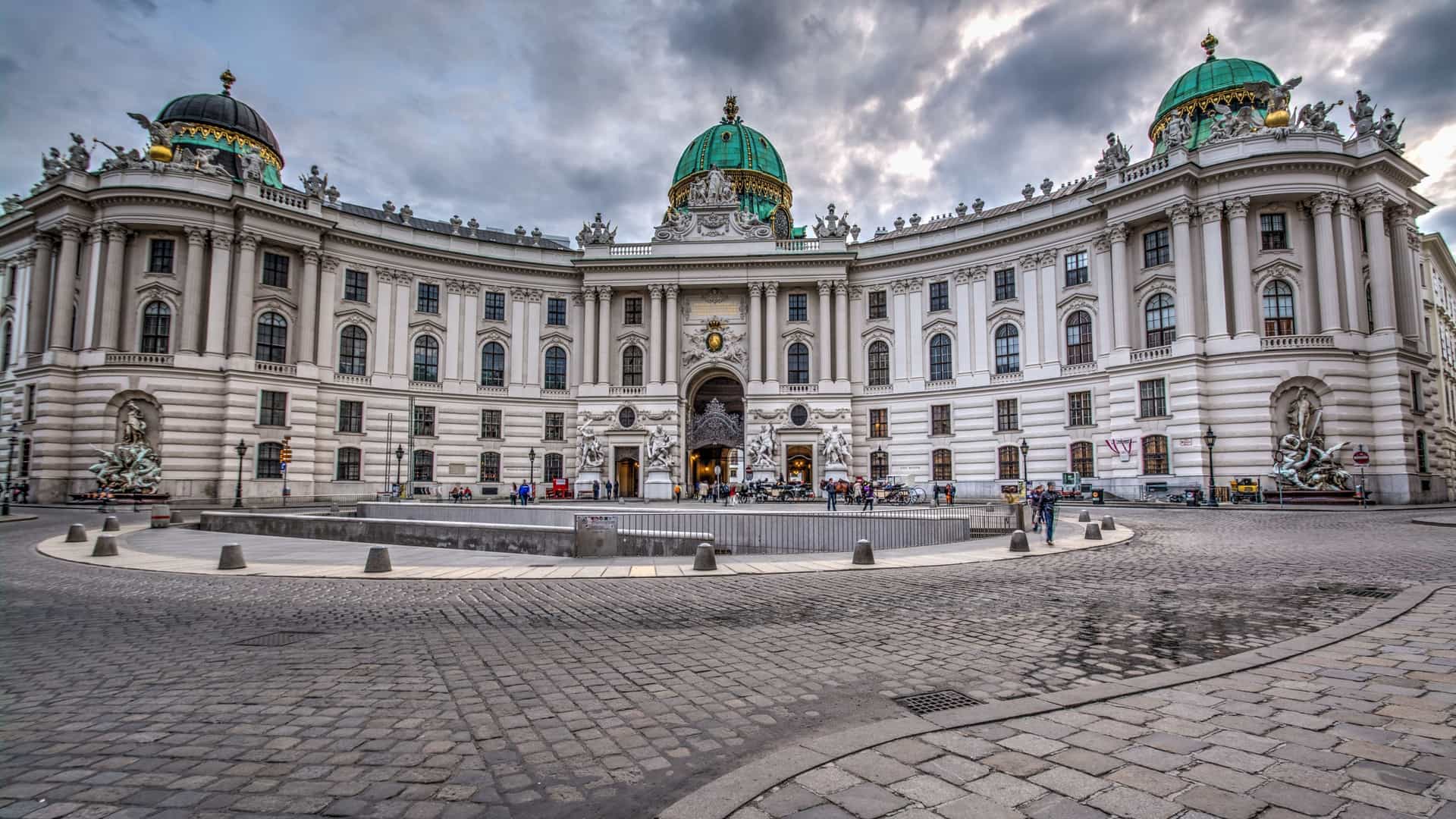 A wide angle view of the Hofburg Palace's Michaelertrakt (St. Michael's Wing) in Vienna, Austria, seen from Michaelerplatz. The grand, Baroque facade, featuring a large domed entrance, is visible, with traditional horse drawn carriages (Fiakers) and tourists occupying the sunny square in the foreground.