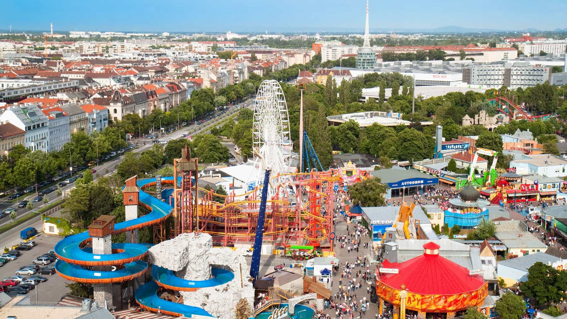 A high angle view of Vienna's Prater amusement park showing the track of a roller coaster and a tall water toboggan ride. The iconic Prater Ferris Wheel is visible in the distance, against a backdrop of trees and the urban landscape of Vienna, all under a sunny sky.