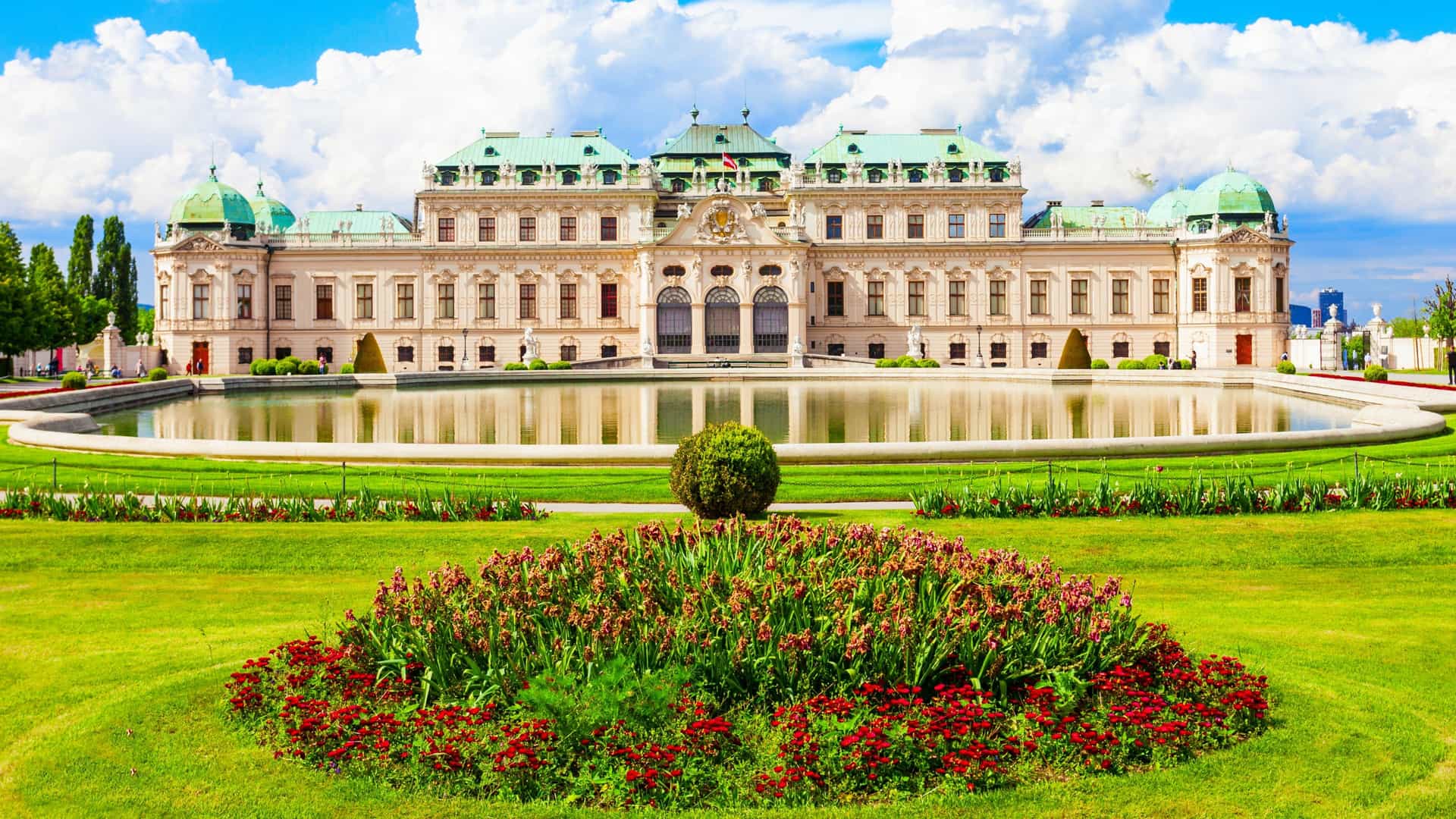 The Upper Belvedere Palace, a grand Baroque structure in Vienna, is viewed across the formal, symmetrical Baroque gardens. The palace's magnificent facade, with its ornate central dome and light colored stone, is reflected in a large, still rectangular reflecting pool, which is flanked by meticulously manicured lawns and hedges.