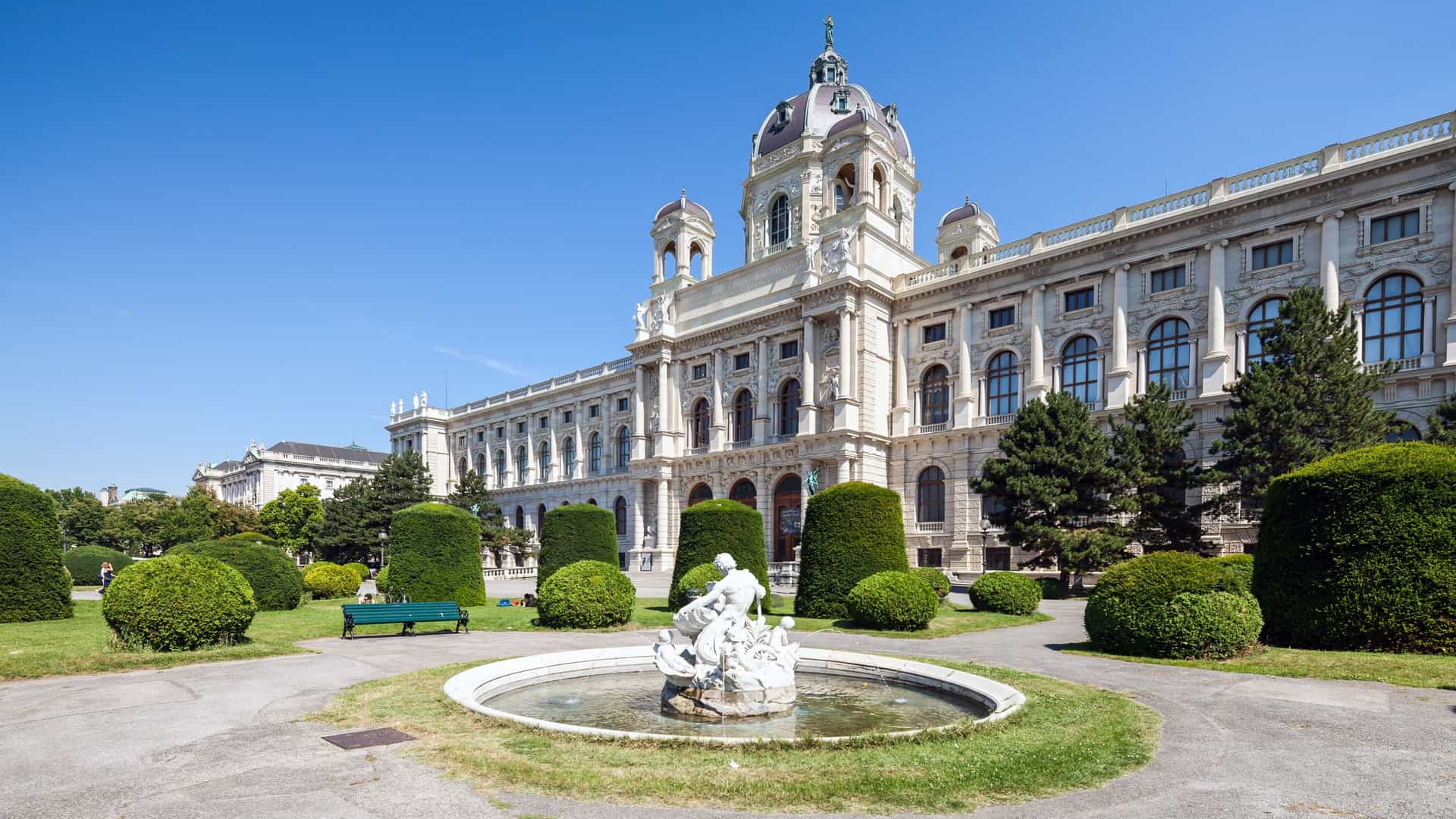 The opulent, historicist facade of the Kunsthistorisches Museum in Vienna is centered under its magnificent octagonal dome, which is crowned with a statue of Pallas Athena. The Renaissance Revival building, constructed of sand colored stone with ornate sculptures and arched windows, stands prominently against a clear blue sky. The image captures the museum's grand, palatial architecture on the Ringstrasse.