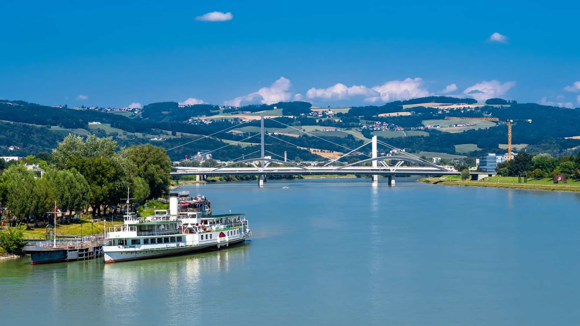 A wide angle view of the Danube River at twilight or dusk, with multiple multi deck river cruise ships docked along the city embankment. The historic buildings of the European city skyline are illuminated, reflecting their lights on the dark water.
