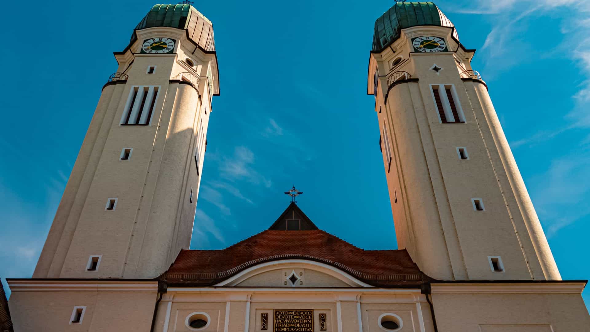 A low angle, upward looking view of the two prominent, light colored bell towers of the Abbey Church (Dreifaltigkeitskirche) at Schweiklberg Abbey (also known as Kloster Vilshofen) in Bavaria, Germany. The towers feature clock faces near their copper domed tops, and the central section of the church's facade with its arched entrance is visible between them, all set against a clear blue sky.