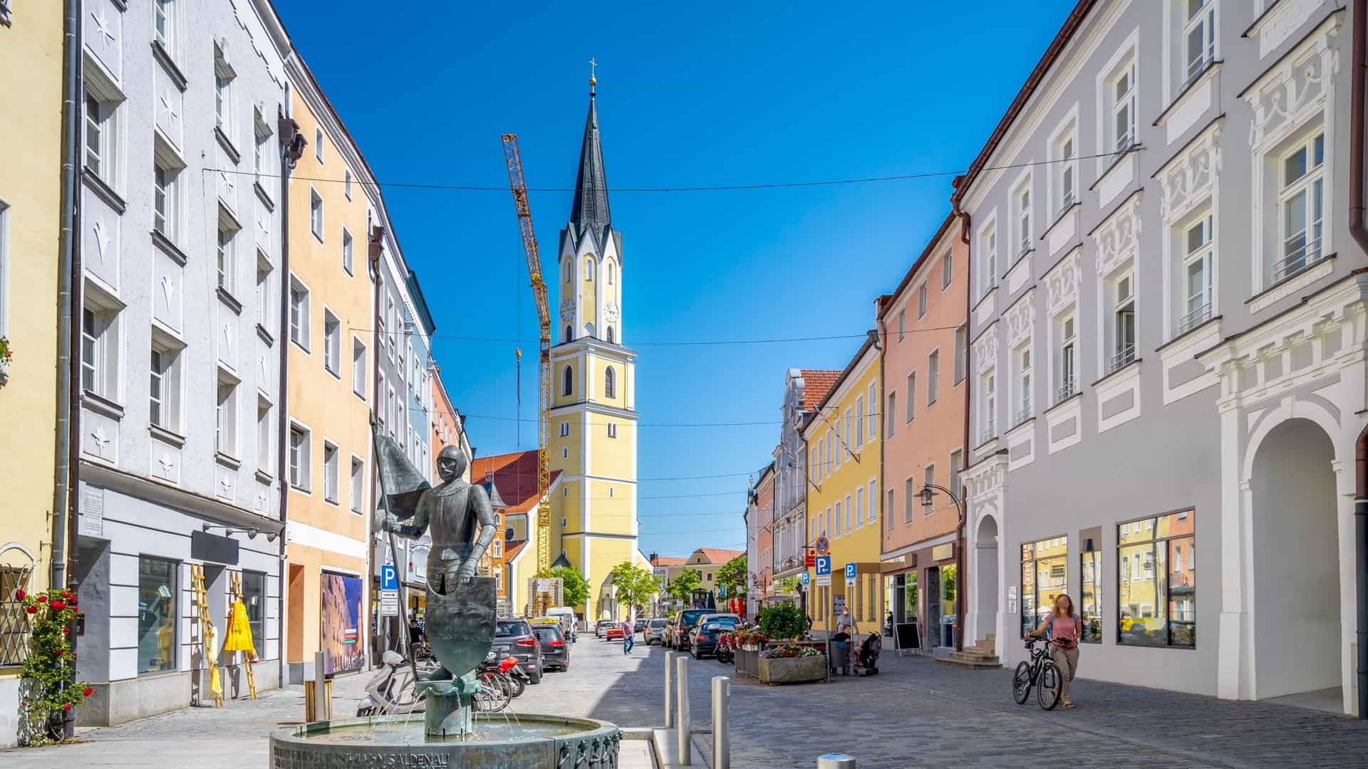 A view down a bustling pedestrian street in Vilshofen's Old Town (Altstadt) in Bavaria, Germany. In the foreground, a bronze fountain with the Ritter Alain (Knight Alain) statue stands. The street is lined with colorful, historic buildings, leading towards the prominent yellow and white spire of the Parish Church of St. John the Baptist (Stadtpfarrkirche St. Johannes der Täufer) in the background. A construction crane is visible behind the church spire.