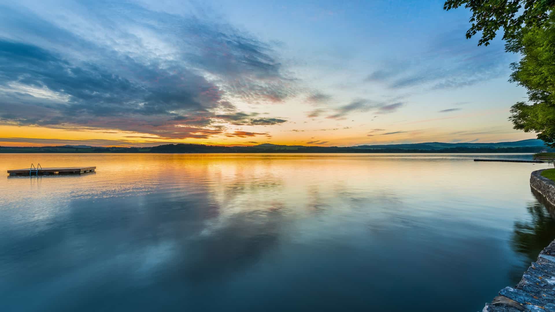 A serene sunset over the reflective surface of Wallersee, a picturesque lake located a short drive from Vilshofen. The sky is filled with dramatic, colorful clouds of blue, orange, and yellow that reflect clearly on the calm, deep blue water. A small, wooden dock floats on the left, and a stone embankment with a dark green tree borders the right foreground.