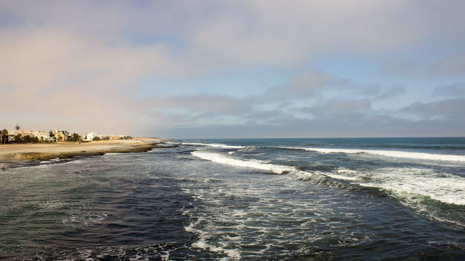 A scenic view of the Langstrand coastline in Namibia. The image features a stretch of beach with sea side property developments and buildings along the shore. The Atlantic Ocean meets the sand under a partly cloudy blue sky.