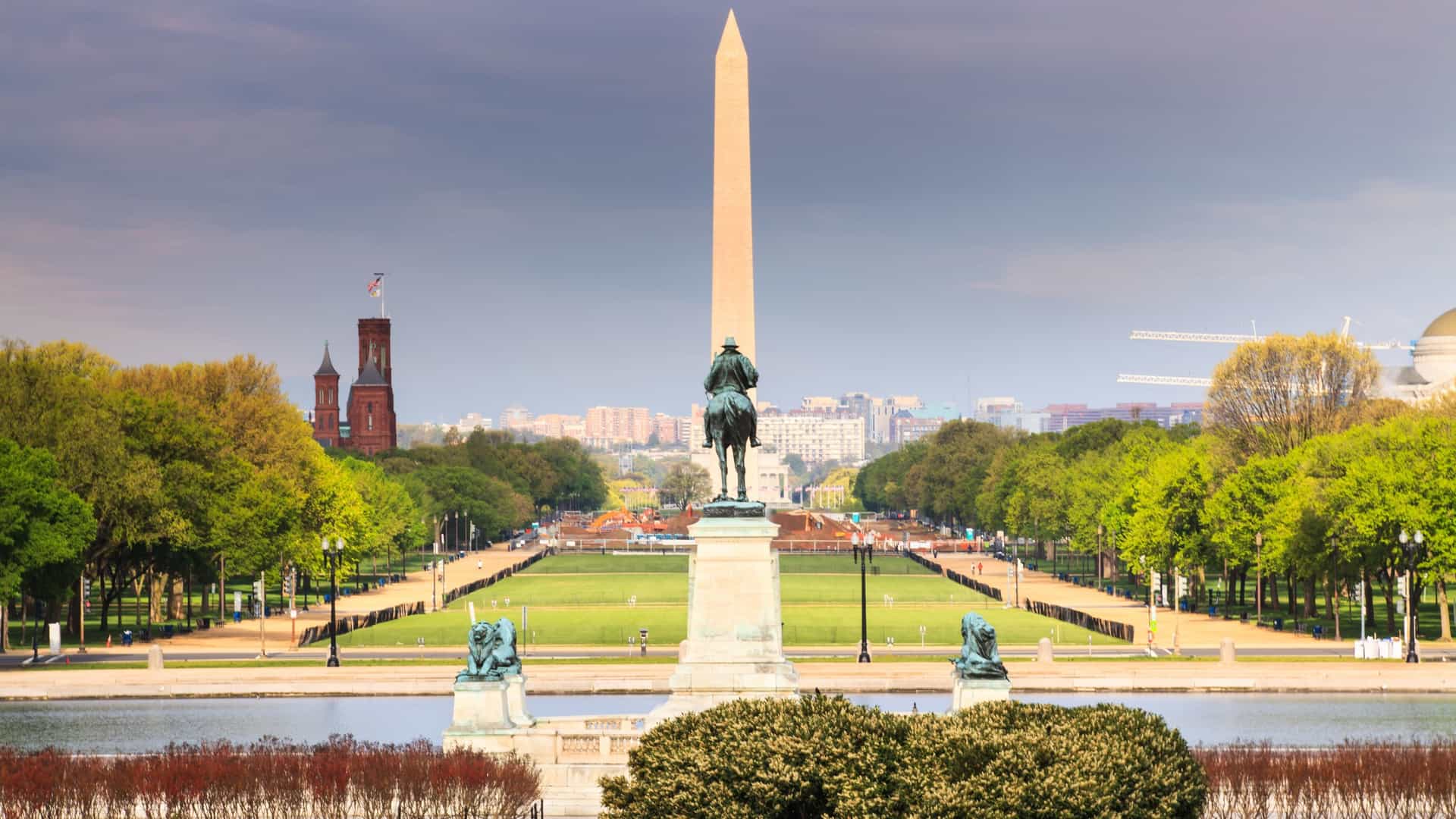 A long, sweeping view of the National Mall in Washington, D.C., taken from the U.S. Capitol Building. The perspective looks westward down the length of the grassy, tree lined Mall, with the Ulysses S. Grant Memorial in the foreground and the prominent Washington Monument visible in the distance.