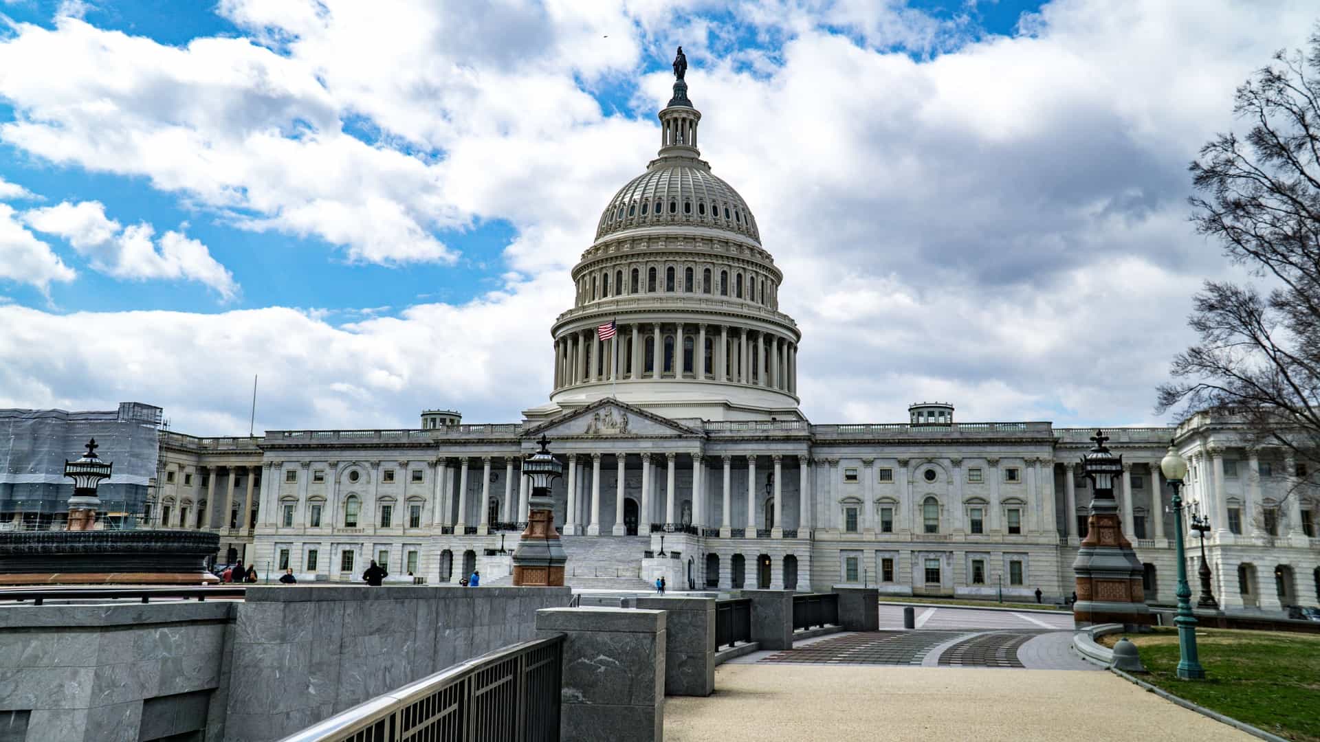 The United States Capitol Building in Washington, D.C., the home of the U.S. Congress. The neoclassical building is topped by its iconic white dome and is set against a blue sky, likely showcasing either the East or West facade with its columns and steps.