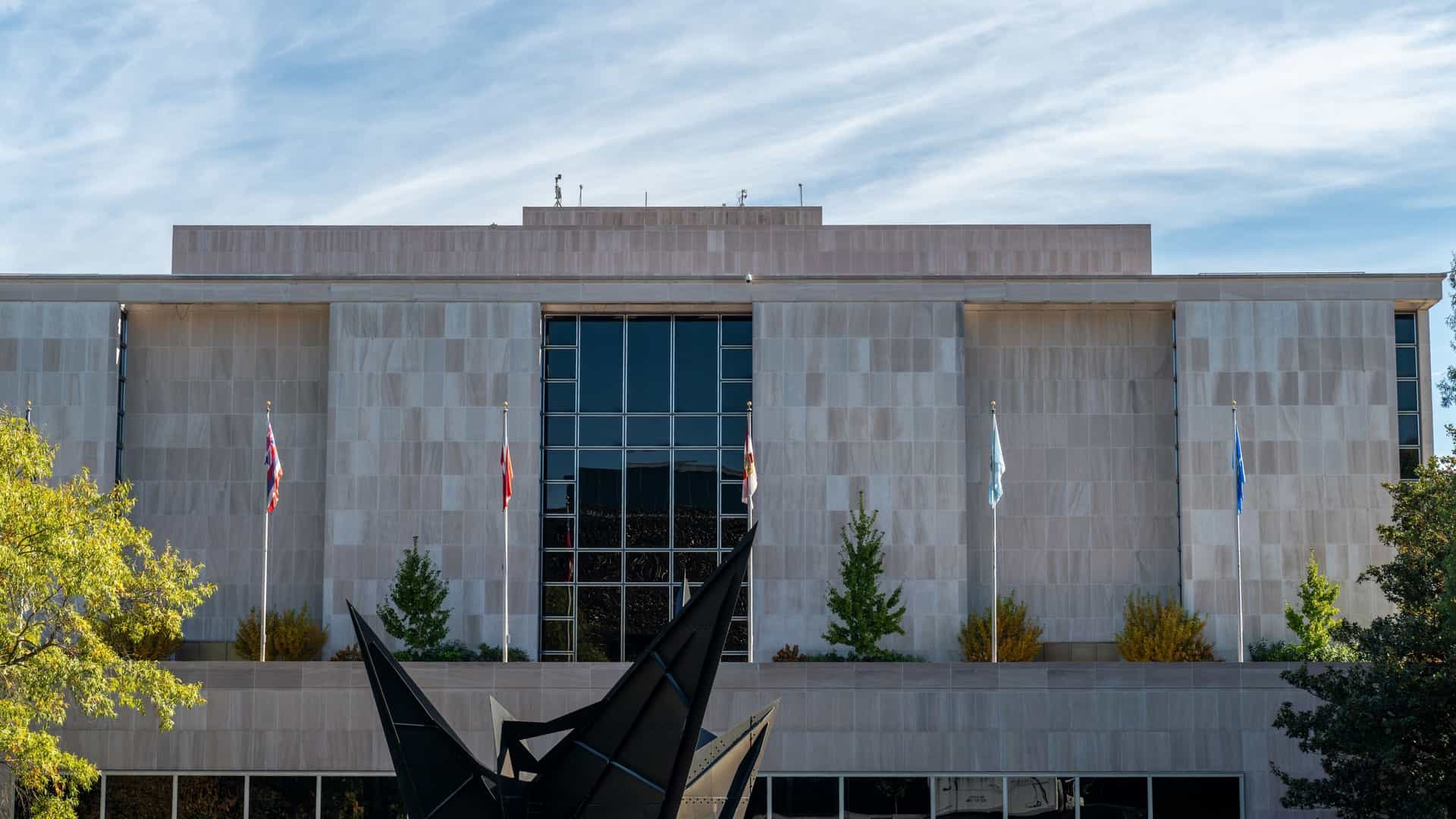 A highly symmetrical and frontal view of the Smithsonian National Air and Space Museum in Washington, D.C. The modern, rectangular building is made of stone and glass, with a central facade and wings extending symmetrically on either side. The architecture features clean lines and a modern, monumental design.