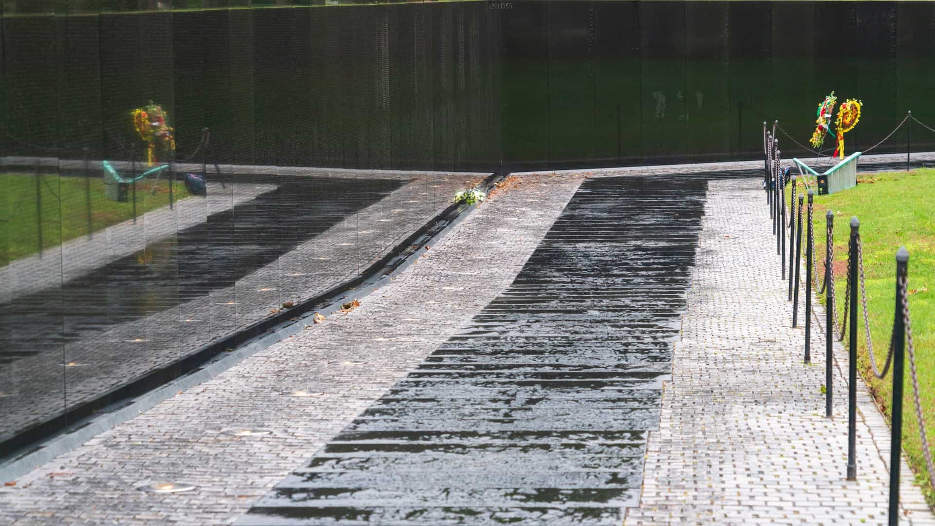 The Vietnam Veterans Memorial in Washington, D.C. The scene features the reflective black granite Wall, engraved with the names of the fallen, meeting the adjacent bronze statue, The Three Servicemen. The statue depicts three figures in military uniform standing guard and gazing towards the Wall, symbolizing their solemn tribute to their comrades.