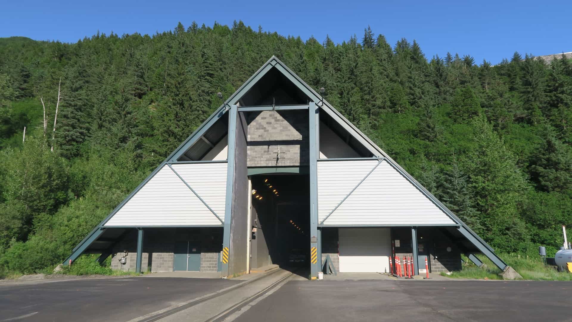The Anton Anderson Memorial Tunnel entrance in Whittier, Alaska. The tunnel's portal is a rugged, A frame structure designed to withstand avalanches, with a large overhead door. The image captures the unique dual use tunnel, which is the longest combined vehicle and railroad tunnel in North America, set against a backdrop of snow capped mountains.