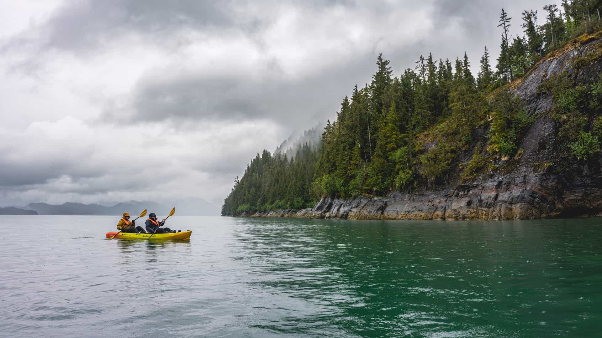 A lone kayaker in a brightly colored boat paddles across tranquil, deep blue water, surrounded by the dramatic, steep sided mountains of an Alaskan fjord. The surface of the water is calm and reflective, mirroring the rugged, tree covered slopes that rise directly from the sea, emphasizing the remote and serene wilderness setting.