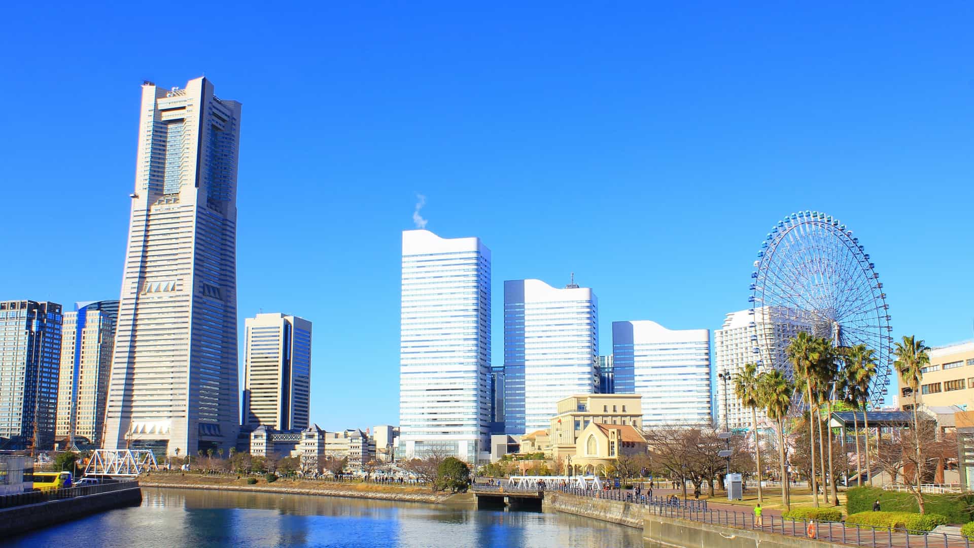 A stunning panoramic view of the Minato Mirai 21 skyline in Yokohama, Japan, likely captured during evening or nighttime. The image prominently features the modern skyscrapers, including the Yokohama Landmark Tower, illuminated against the dark sky. In the foreground or mid ground, the colorful, brightly lit Cosmo Clock 21 Ferris wheel is visible, reflecting in the harbor water.
