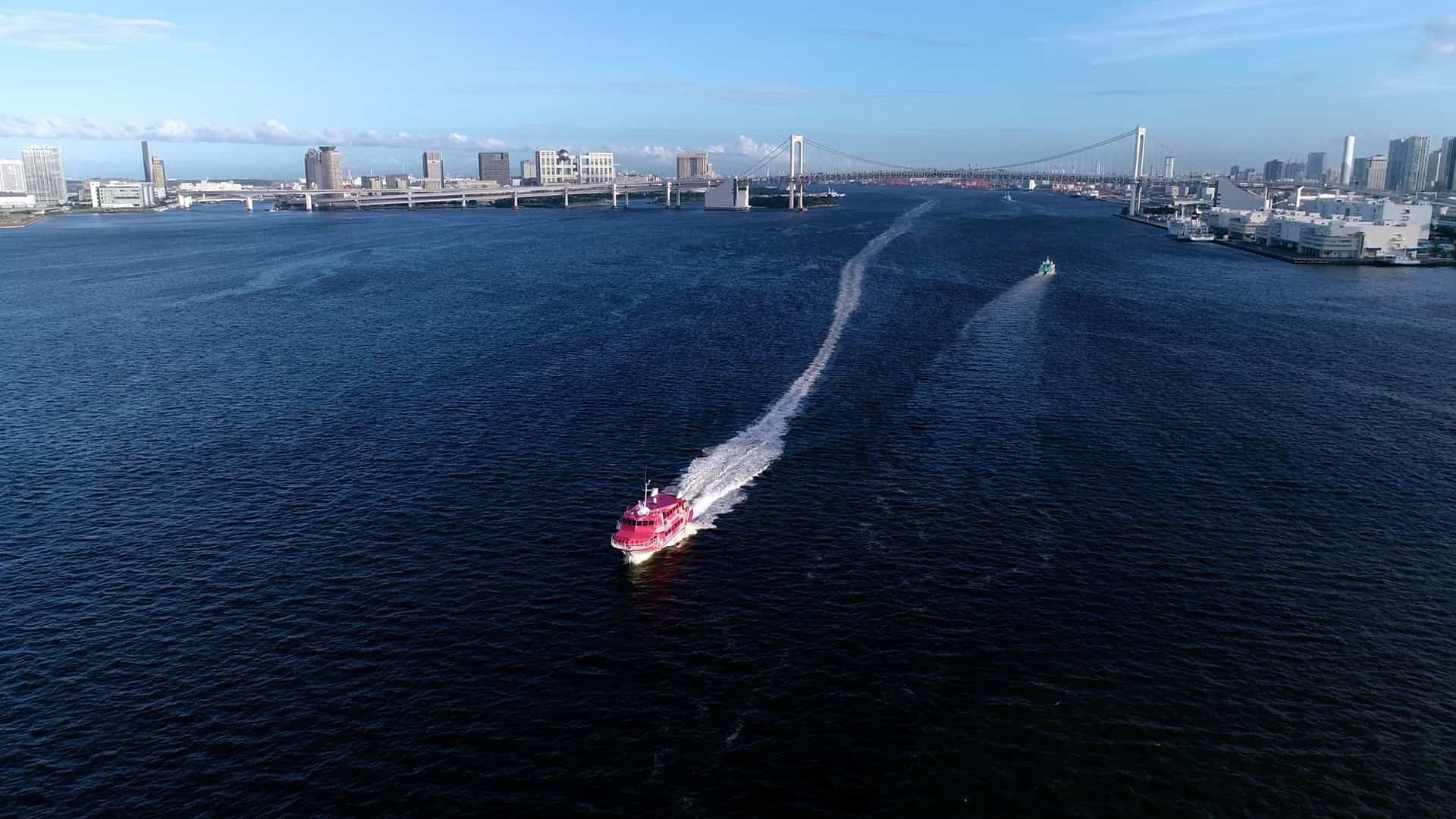 An aerial, high angle view of a boat, likely a pleasure or cruise vessel, sailing on the deep blue waters of Tokyo Bay, Japan. The background is dominated by the dense, modern skyline of the Tokyo waterfront, which may include landmarks like the Rainbow Bridge or the Tokyo Tower in the distance.