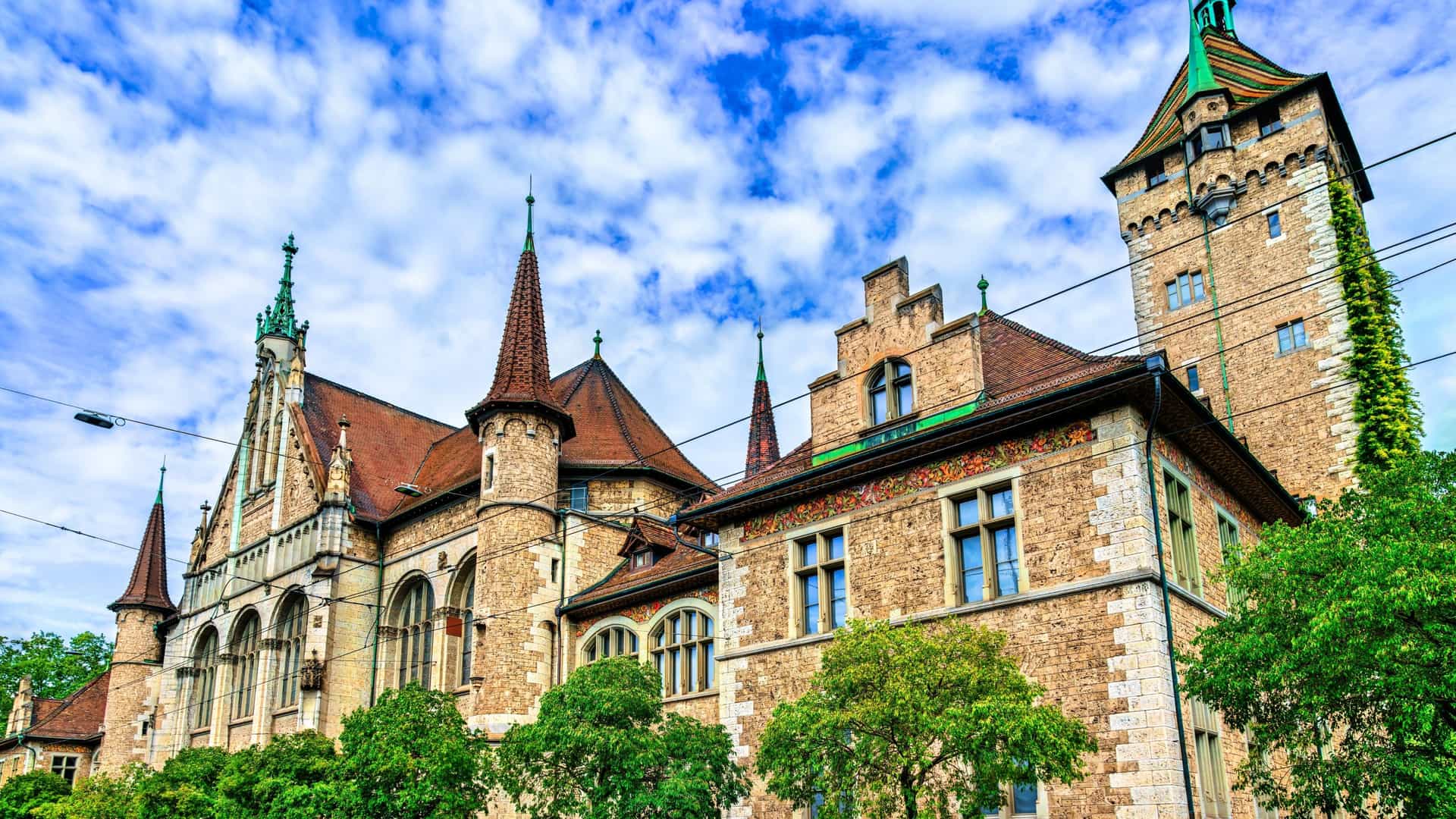 An exterior view of the Swiss National Museum (Landesmuseum) in Zurich, Switzerland, featuring its historicist architecture designed by Gustav Gull, which resembles a French Renaissance chateau with multiple turrets and towers. The image captures the impressive façade of the older section of the museum, situated next to the modern wing, with a clear sky overhead.