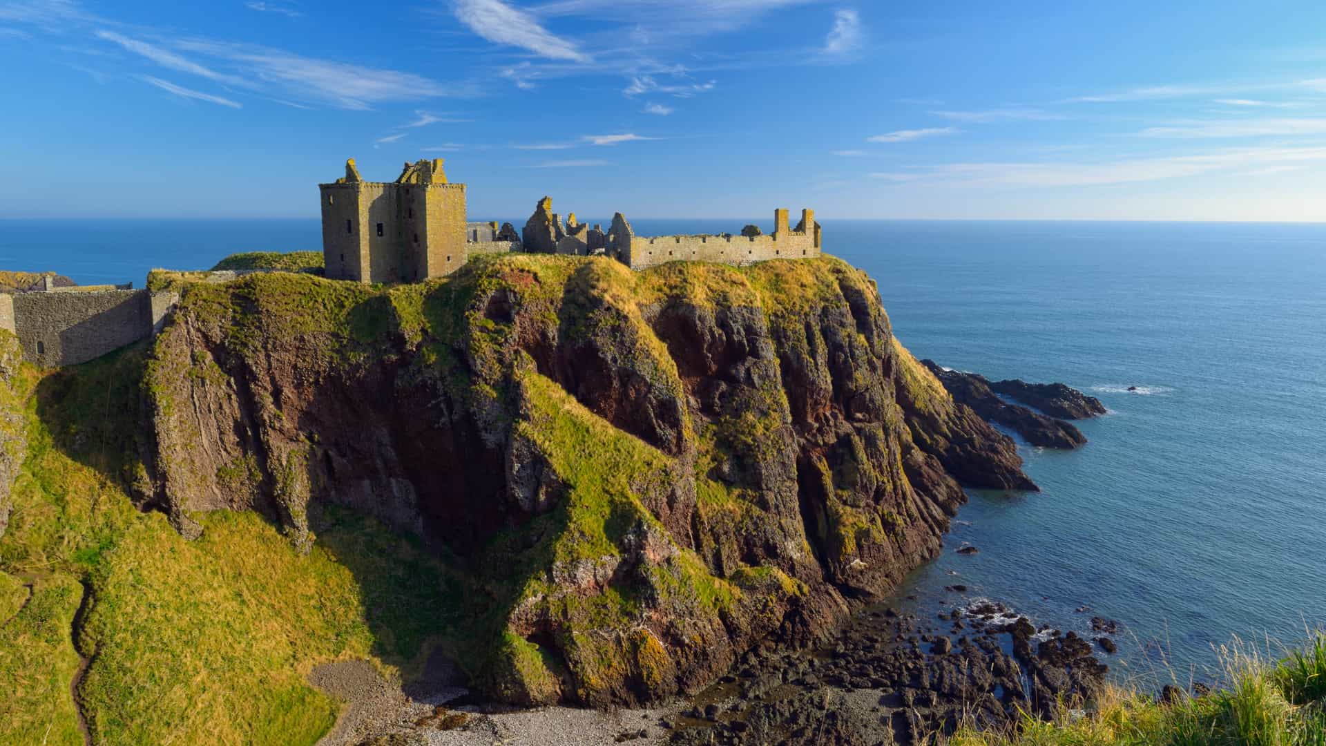 A dramatic coastal scene at Dunluce Castle featuring medieval stone ruins perched on a steep, grassy cliff above the ocean, with rugged rock formations below and waves breaking along the shoreline under a bright blue sky with wispy clouds.