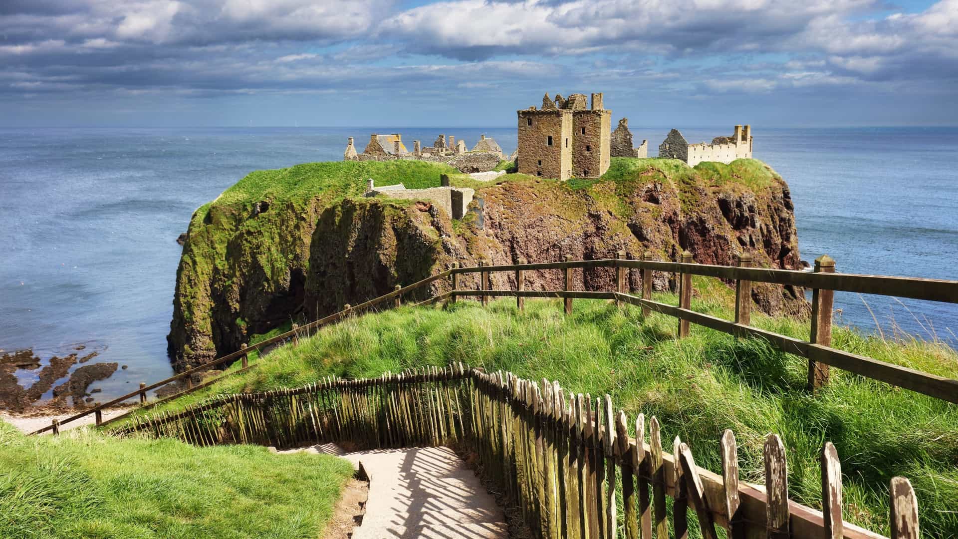An iconic view of the ruined medieval Dunnottar Castle perched on a dramatic cliffside in Am Baile, Scotland, featuring a winding coastal path with wooden fences overlooking the North Sea under a cloudy sky.