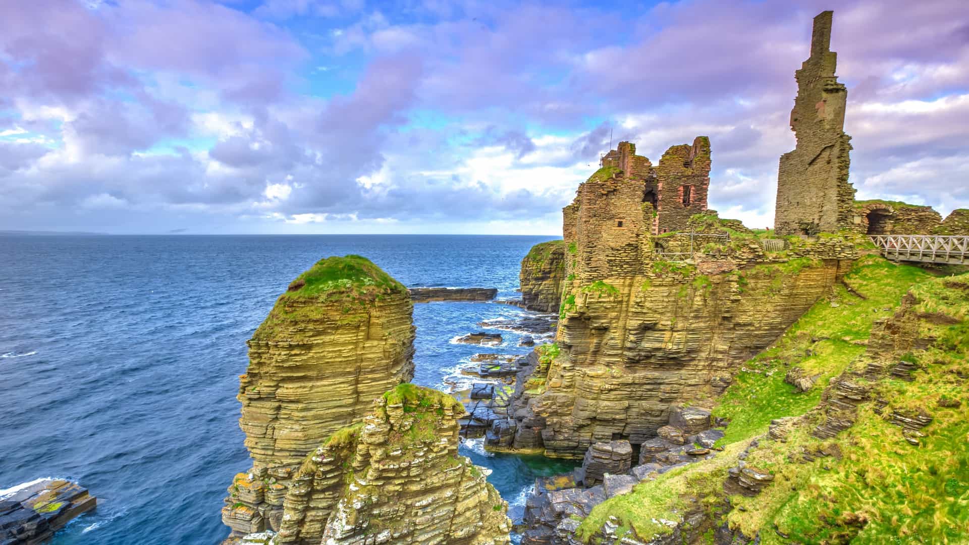 A breathtaking view of the crumbling ruins of Castle Sinclair Girnigoe near Baile Mòr, Scotland, perched precariously on high sandstone cliffs and sea stacks overlooking the vibrant blue waters of Sinclair's Bay under a dramatic, colorful sky.