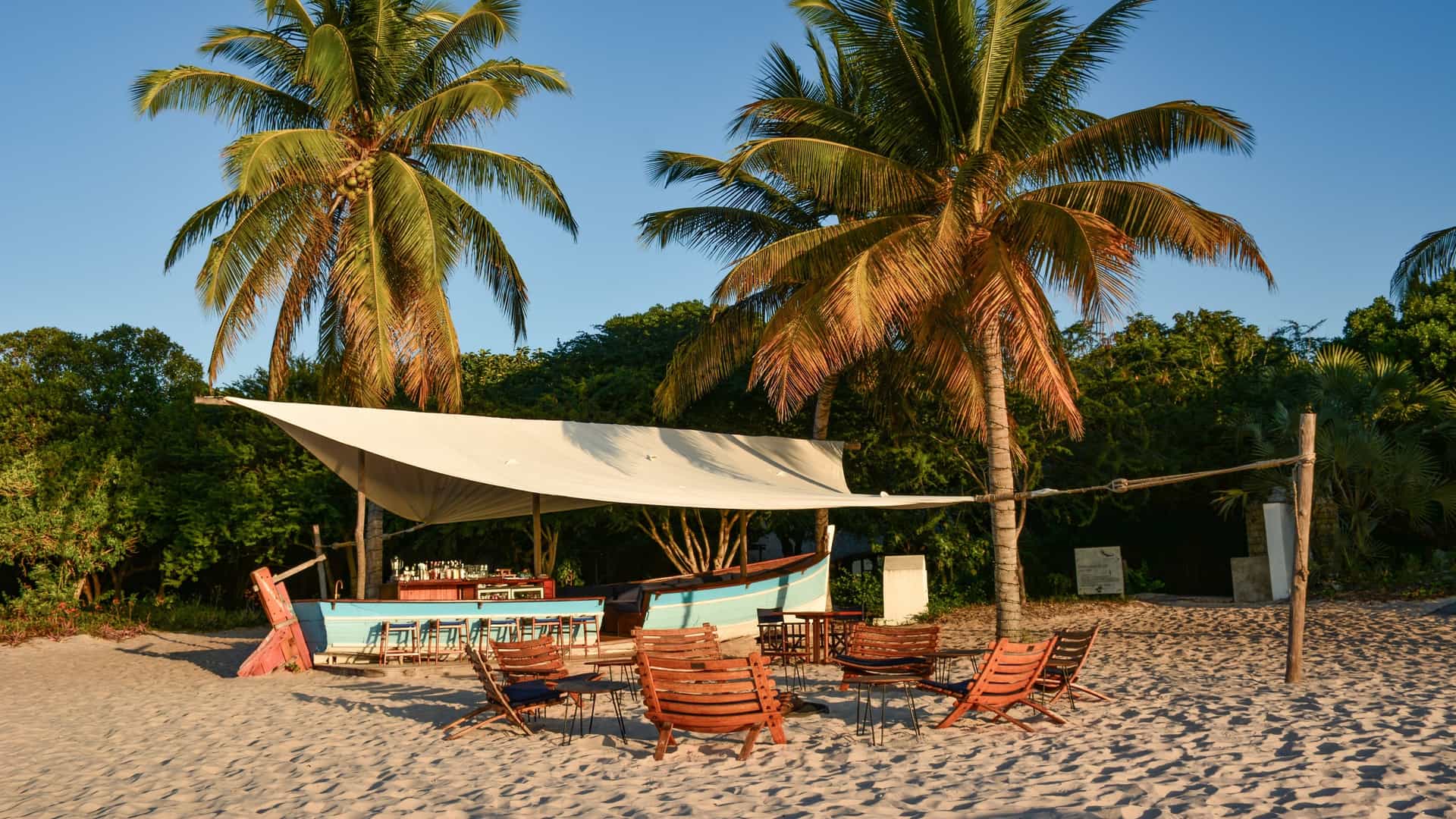 An inviting outdoor beach bar on Benguerra Island, Mozambique, featuring a rustic blue dhow boat converted into a bar under a white canopy, surrounded by wooden lounge chairs on white sand and lush tropical palm trees.
