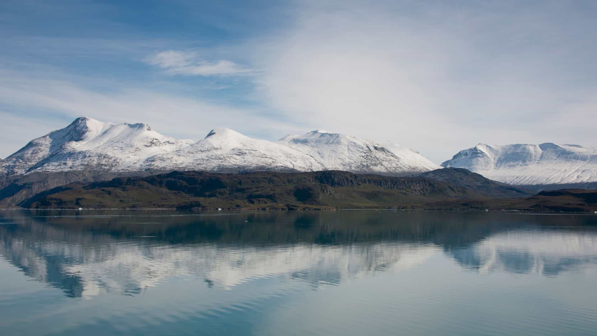 A stunning landscape view of the snow-capped mountains and icy fjords near Brattahlid, Greenland (part of the Kingdom of Denmark), featuring the calm, reflective waters of the Tunulliarfik Fjord under a bright, clear sky.