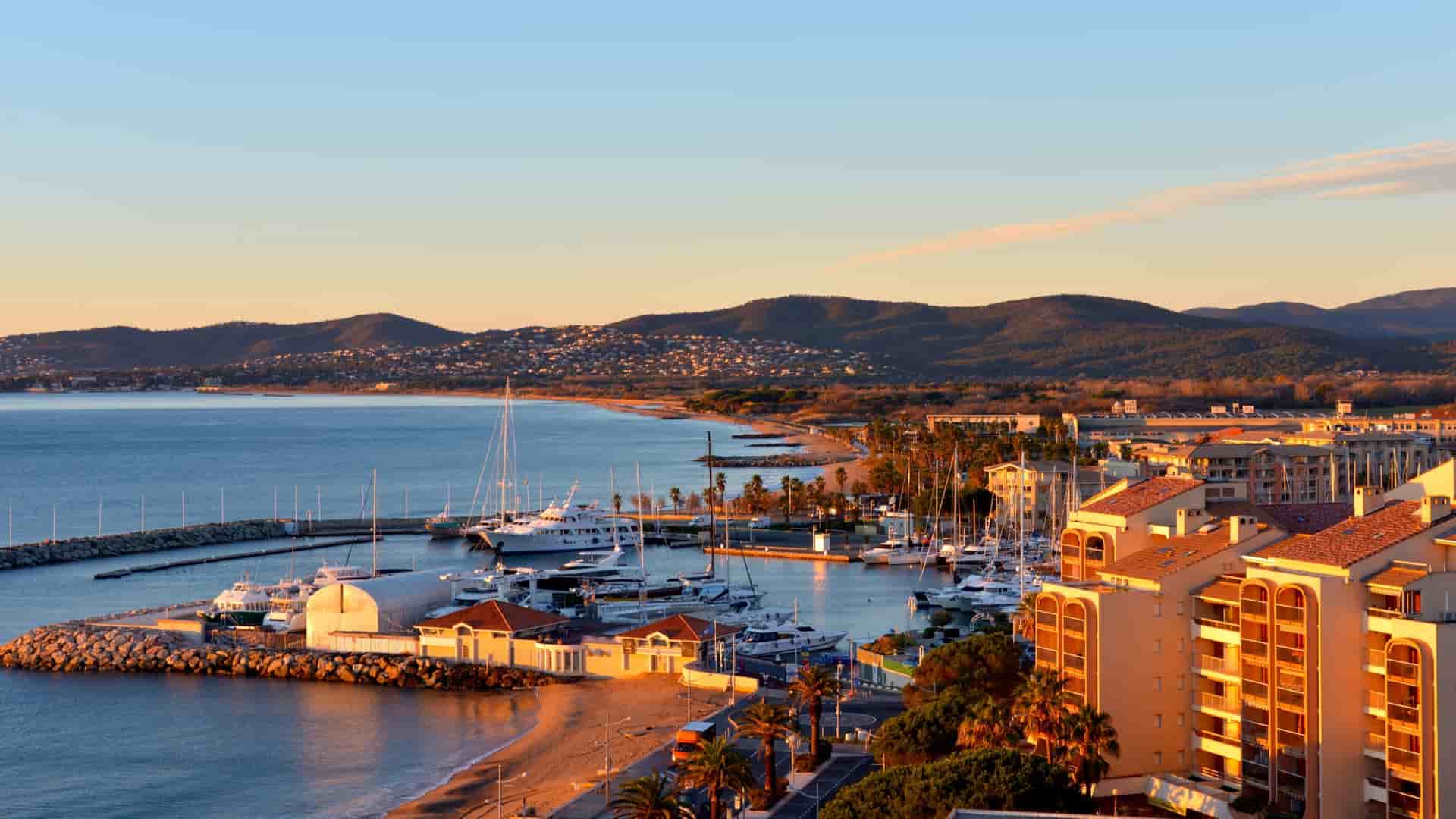 A golden hour view of the Port Fréjus marina in Fréjus, France, featuring luxury yachts moored in the harbor, Mediterranean-style apartment buildings along the coast, and a sweeping sandy beach stretching toward the distant Esterel Mountains under a soft evening sky.