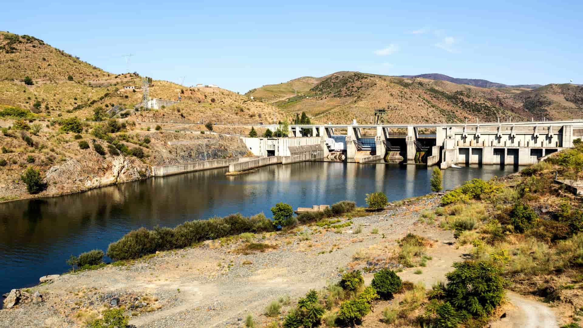 A large concrete dam near Pocinho, Portugal, spanning a calm reservoir in a rugged, semi‑arid landscape. Rocky hills with sparse vegetation rise around the structure, and spillway gates line the front of the dam. Clear blue skies highlight the contrast between the engineered waterway and the natural terrain.