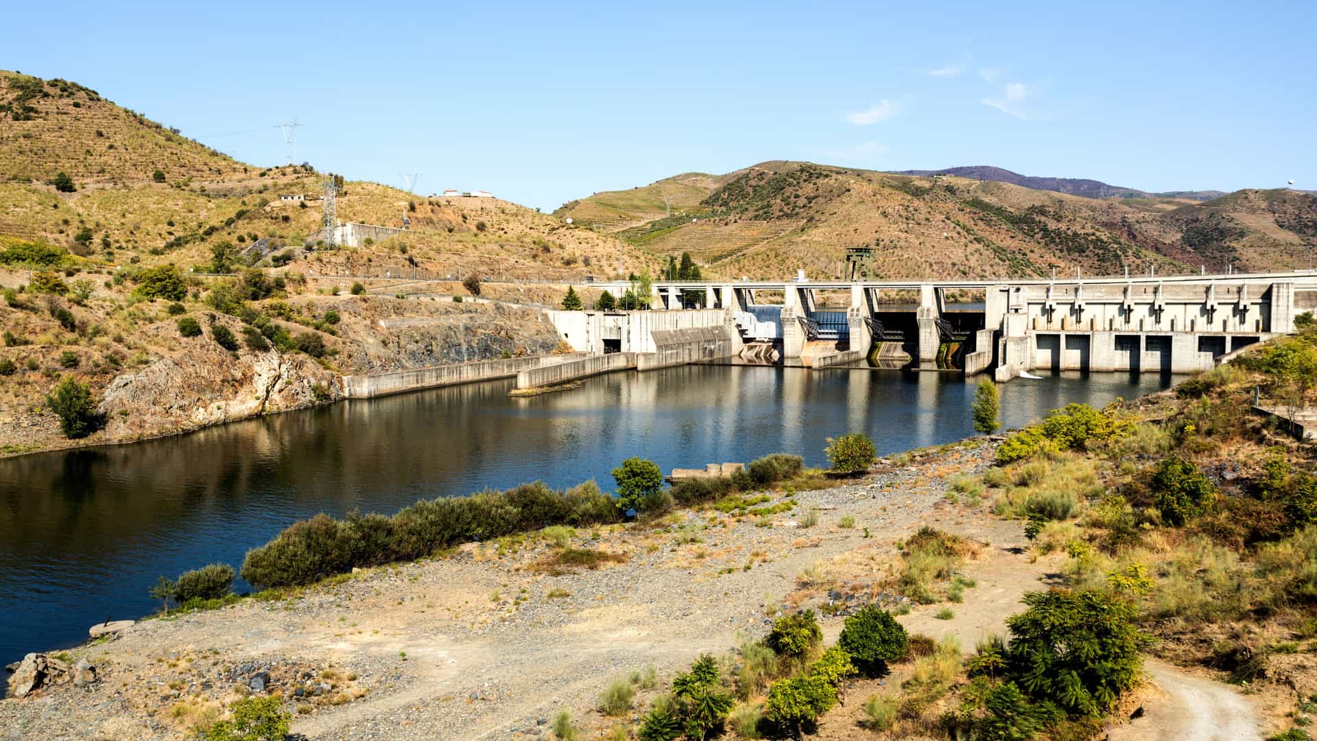 A large concrete dam near Pocinho, Portugal, spanning a calm reservoir in a rugged, semi‑arid landscape. Rocky hills with sparse vegetation rise around the structure, and spillway gates line the front of the dam. Clear blue skies highlight the contrast between the engineered waterway and the natural terrain.