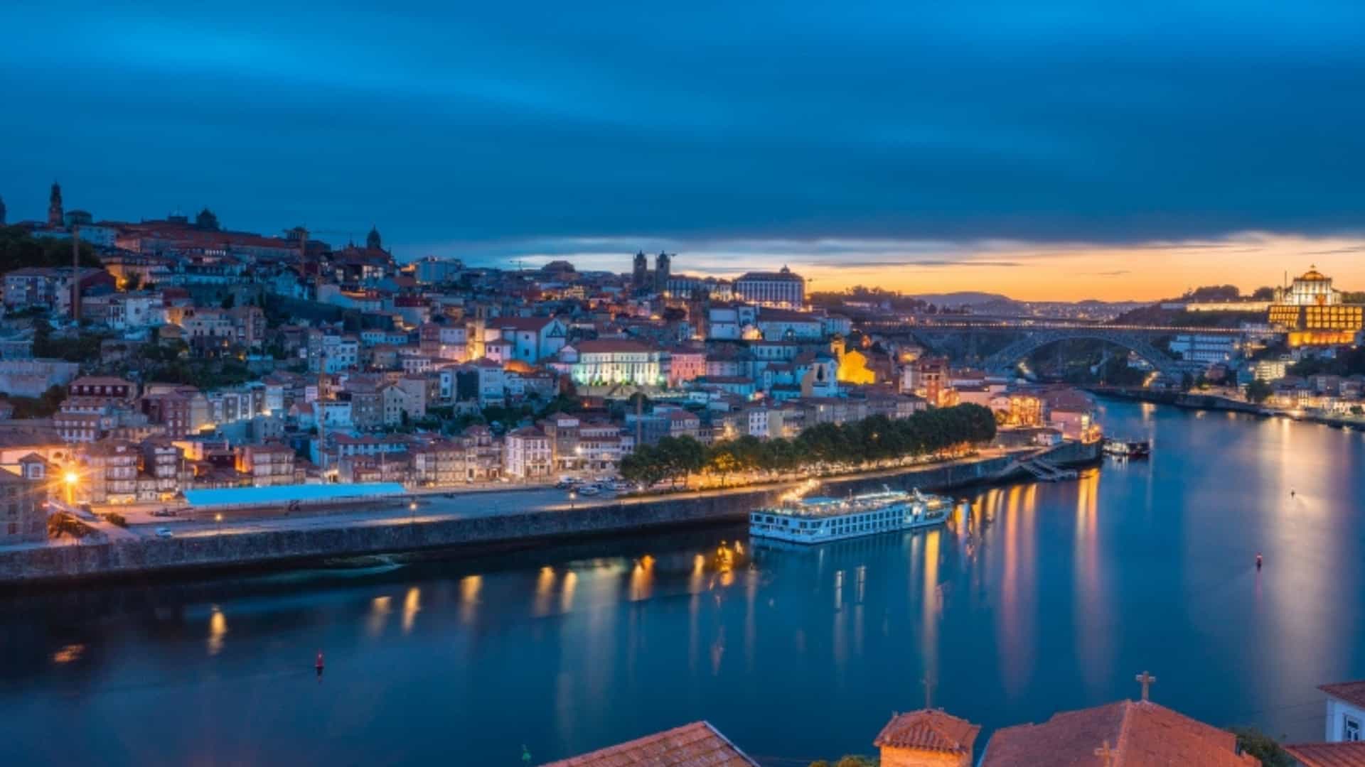 An evening panoramic view of the city of Porto, Portugal, along the Douro River. The city's colorful historic buildings are brightly lit, reflecting on the smooth surface of the river. A long, passenger river cruise boat is docked on the riverbank. In the distance, a bridge spans the river. The sky is a deep blue and orange, marking the transition from sunset to night. The image beautifully captures the illuminated cityscape and the bustling river life of the Douro region.