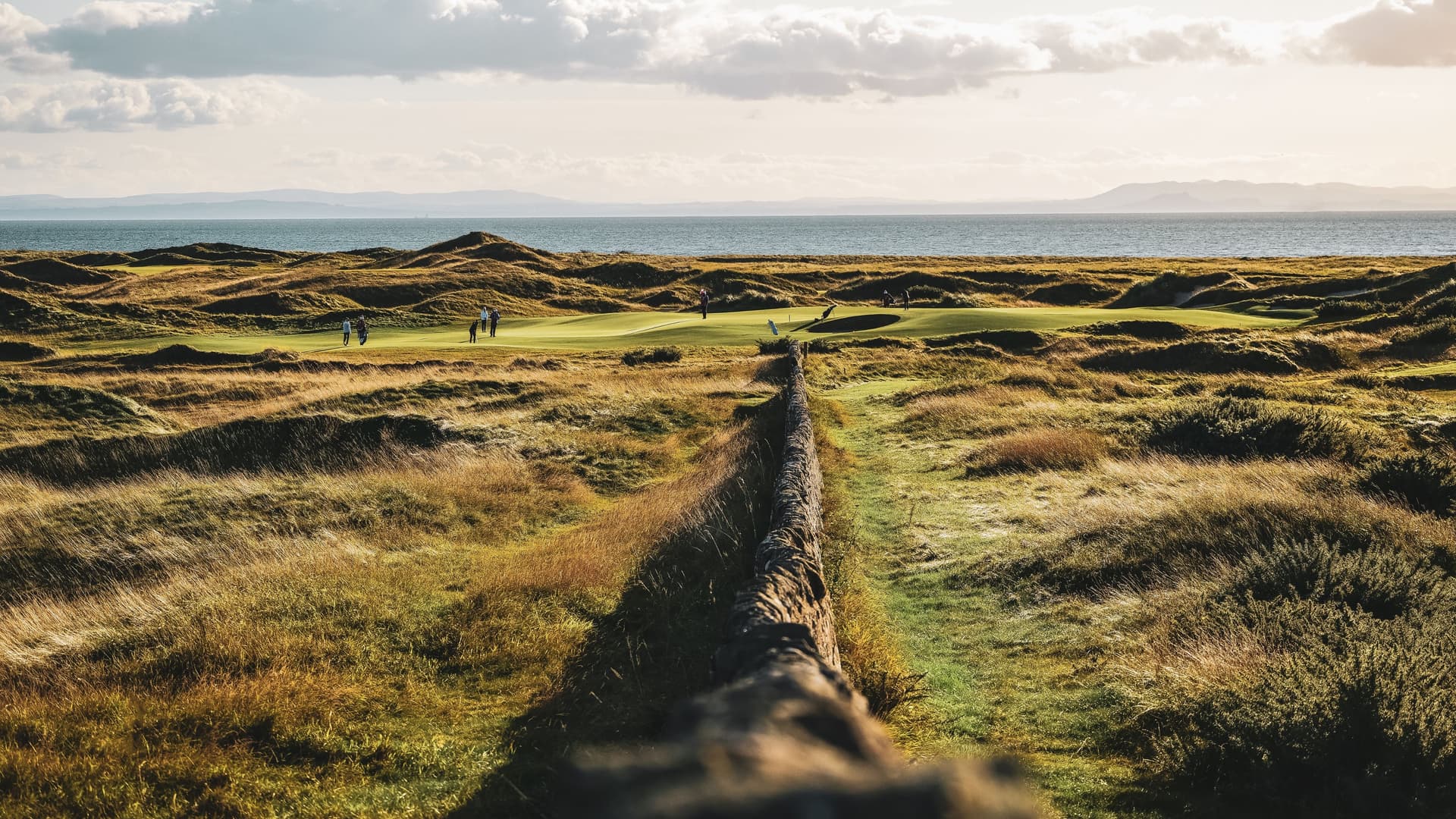 A historic stone wall running through the middle of the rugged, golden grass dunes of Dumbarnie Links in Scotland, with golfers on a green next to the Firth of Forth coastline.