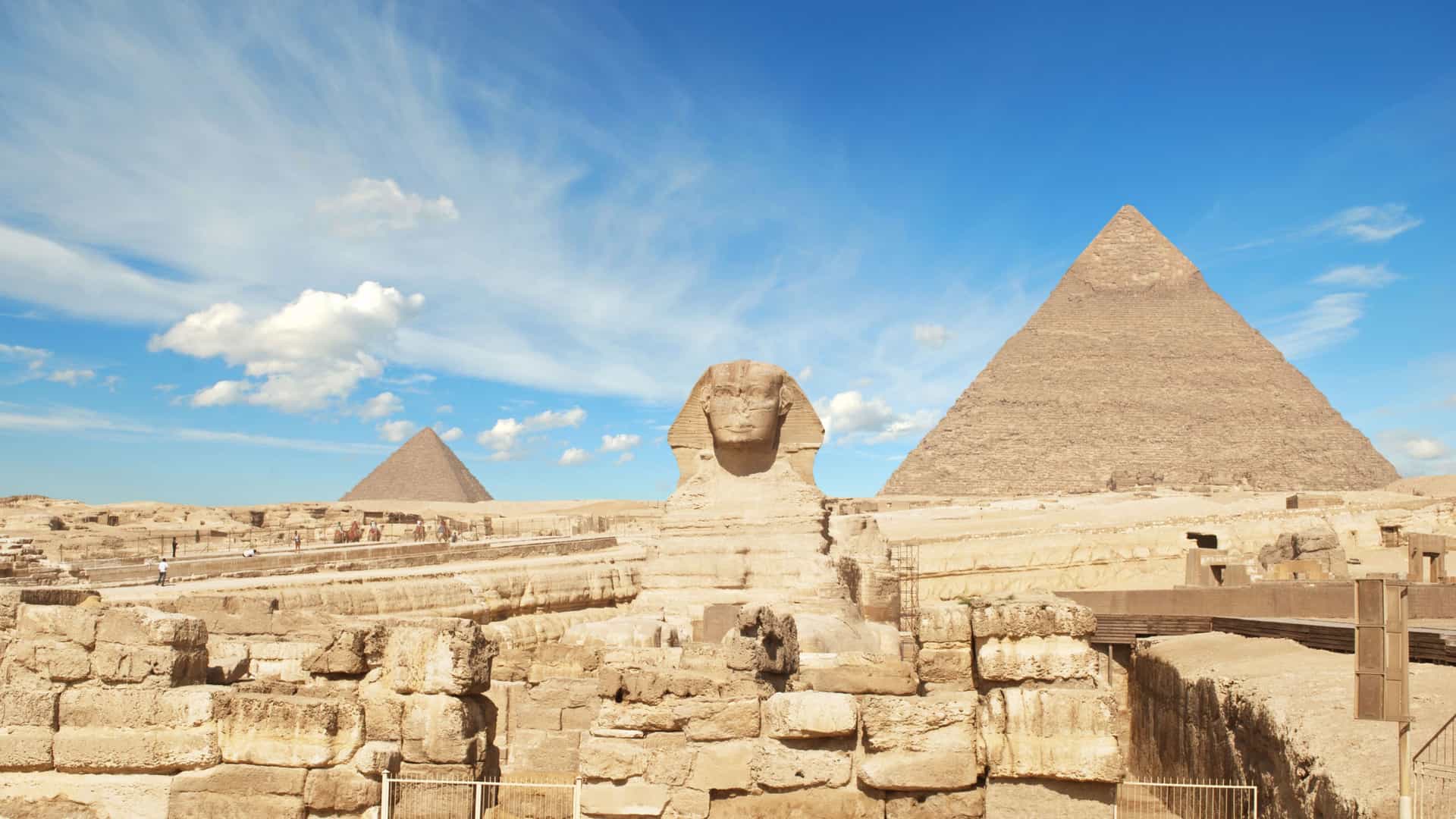 An iconic view of the ancient Giza Plateau in Egypt, a key site along the Nile River. In the center, the Great Sphinx with its human head and lion's body faces to the left. The Sphinx is surrounded by a foreground of large, excavated stone blocks and structures. In the background, the massive, sandy-colored pyramids of Giza rise under a bright, blue sky with wispy clouds. The image captures the historical grandeur and desert landscape of the Egyptian region.