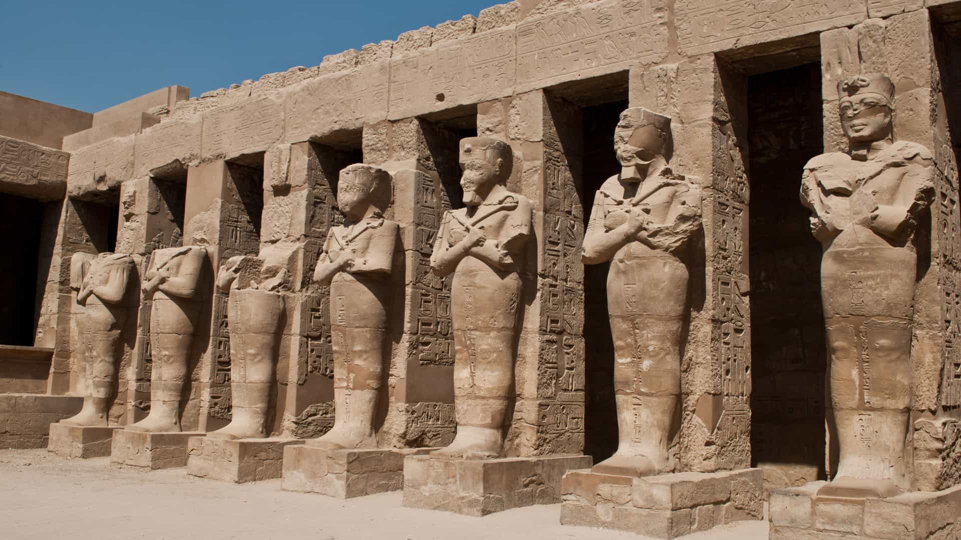 A row of ancient pharaoh statues stands at the entrance of the Karnak Temple in Luxor, Egypt, a historical site located along the Nile River. Each statue is carved from stone, with some appearing weathered and partially damaged over time. The statues are positioned in front of a stone wall covered with intricate hieroglyphs. The ground is sandy, and the sky is a clear, bright blue. The image captures the impressive scale and rich history of ancient Egyptian architecture and art.