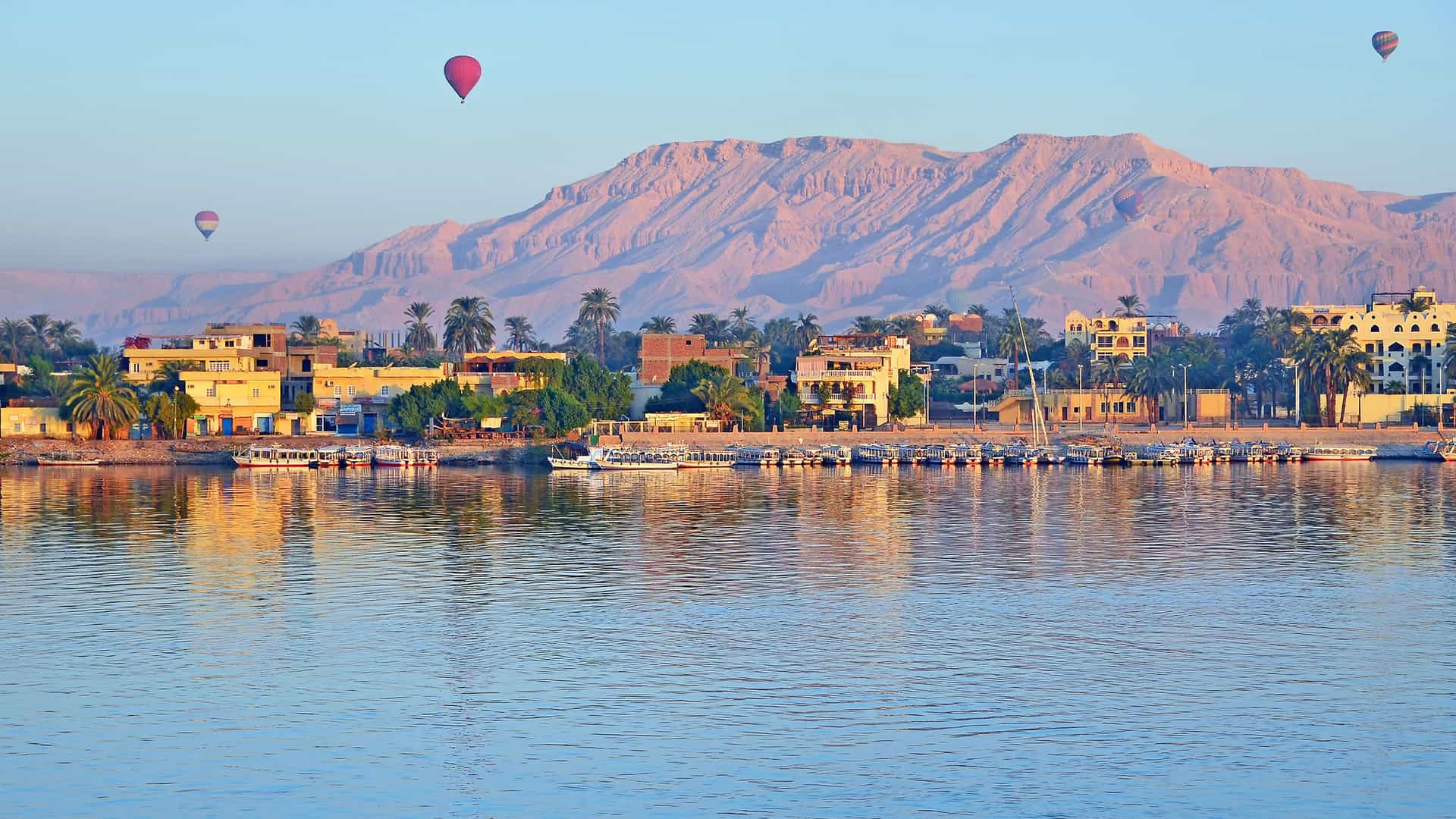 A serene morning view of the Nile River Valley in Egypt, a popular spot for hot air balloon rides. In the foreground, the calm, blue water of the Nile reflects the golden light of the sunrise. On the riverbank, a line of buildings and palm trees stands in front of a large, reddish-brown mountain range. Several colorful hot air balloons float peacefully in the pale blue sky above the mountains, highlighting a popular tourist activity in the Egyptian region.